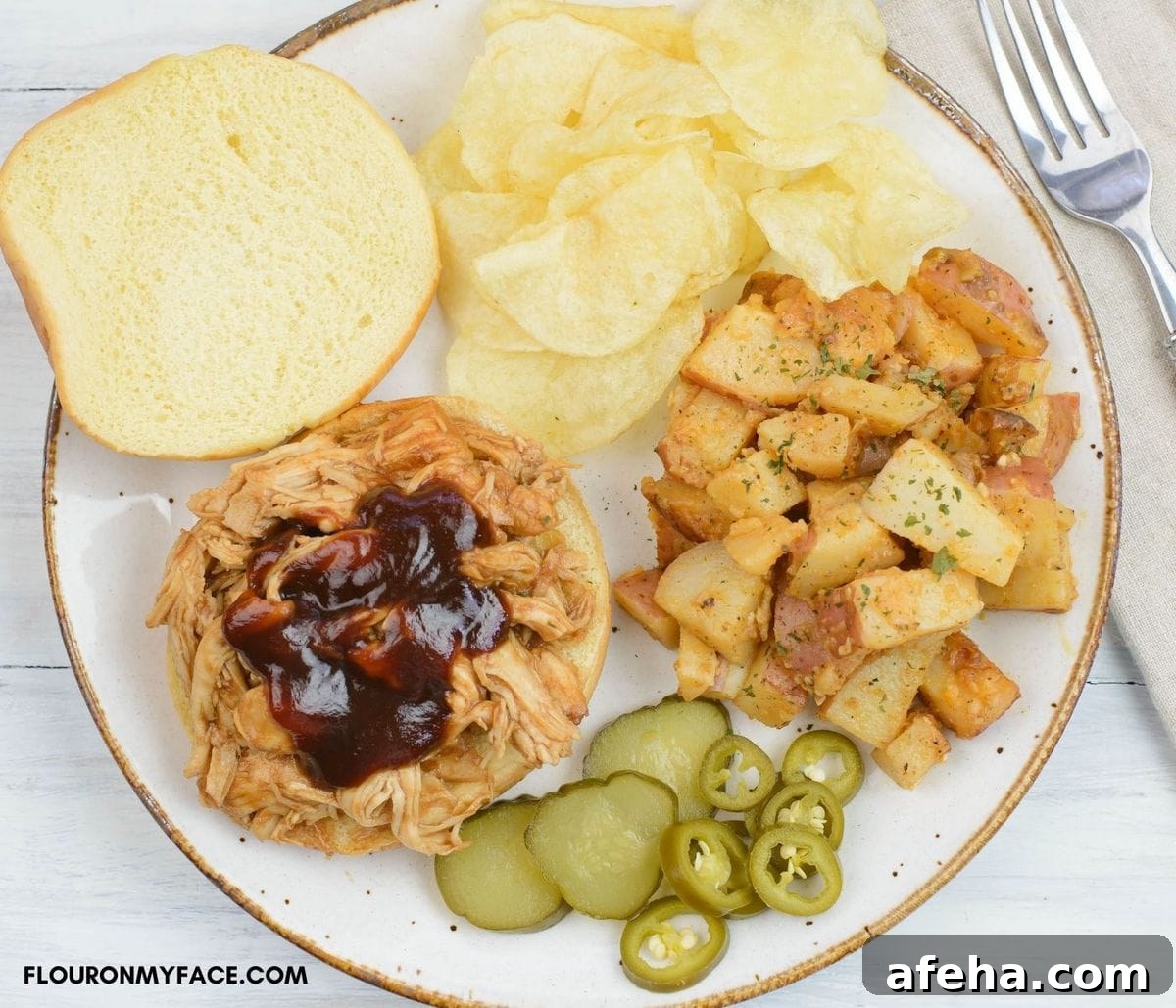 Opend pulled chicken sandwich with barbecue sauce, sides, chips, and slice pickles and jalapeno peppers on a dinner plate.