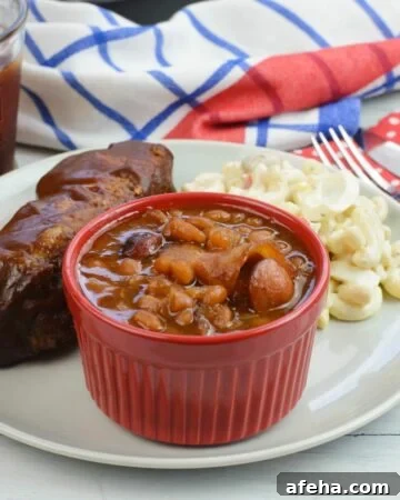 A small bowl filled with baked beans on a plate with ribs.