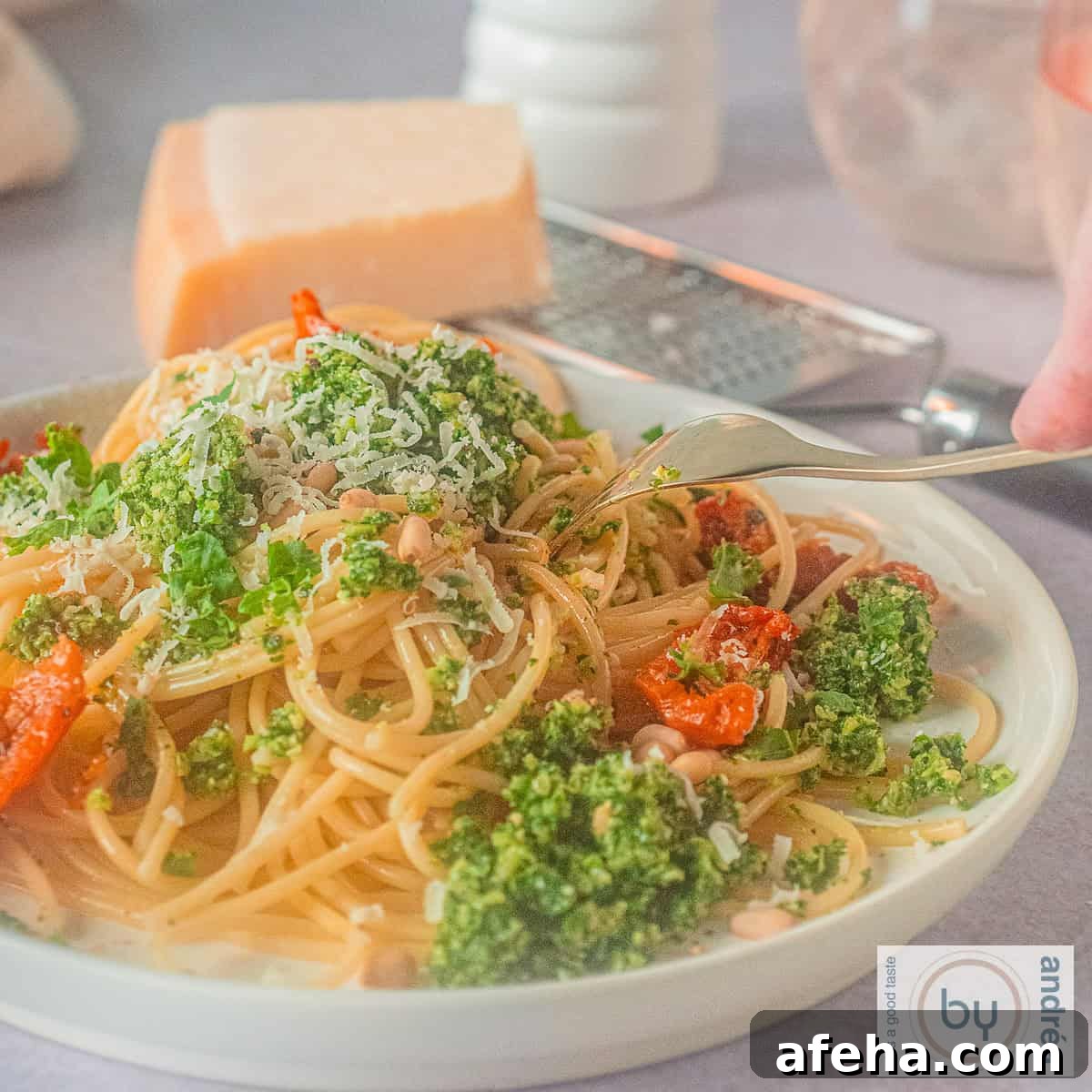 A square photo of a white plate with spaghetti, kale pesto and sun-dried tomatoes. A fork is poking the plate.