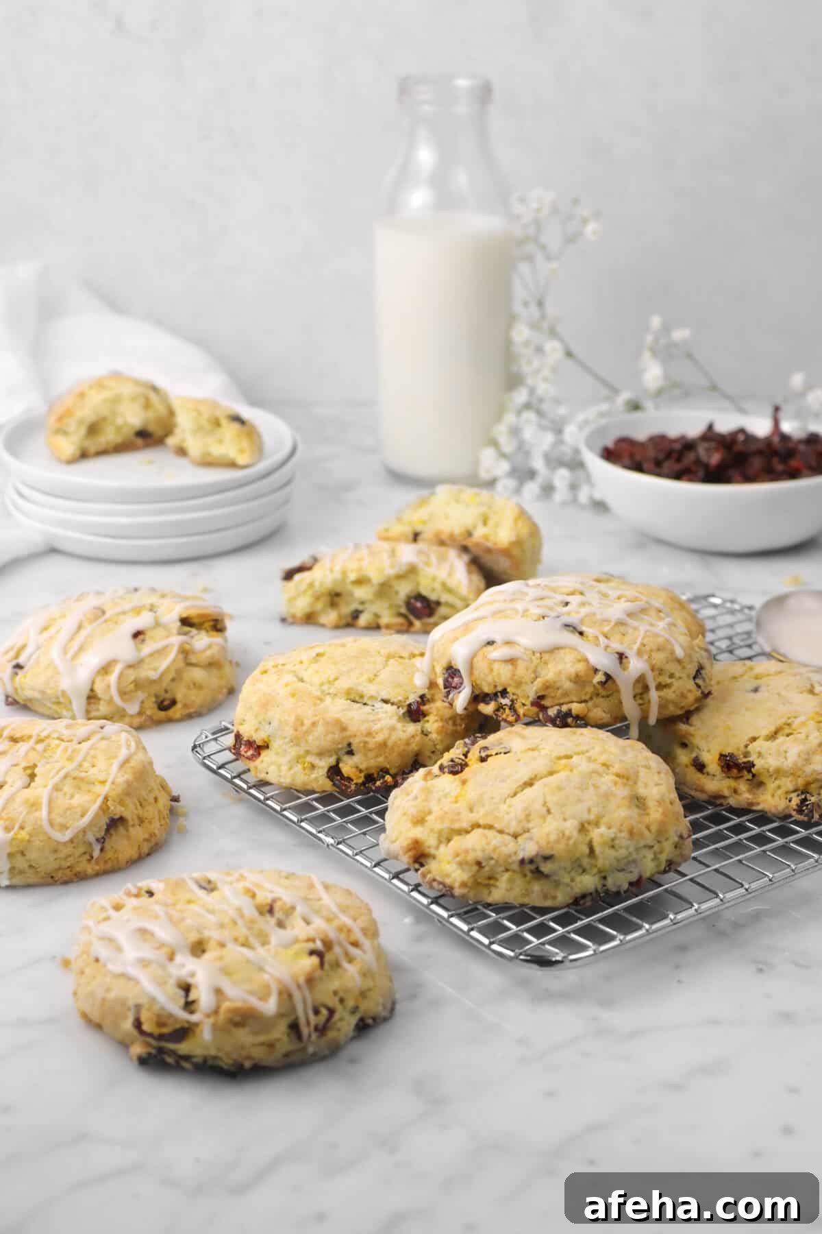 four scones on a cooling rack with three beside, a glass of milk, bowl of cranberries, and flowers behind