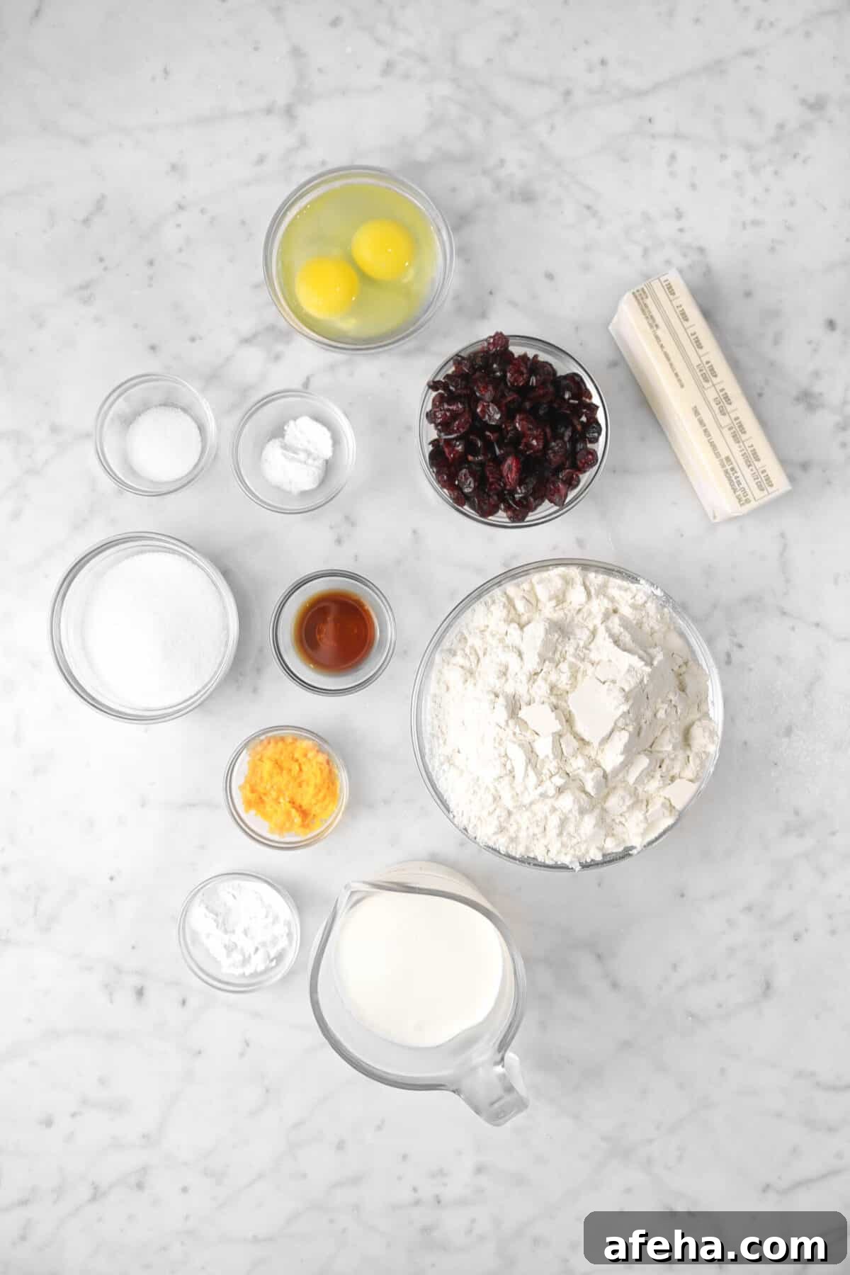 eggs, dried cranberries, butter, baking powder, salt, sugar, vanilla, orange zest, baking soda, milk, and flower on a marble counter