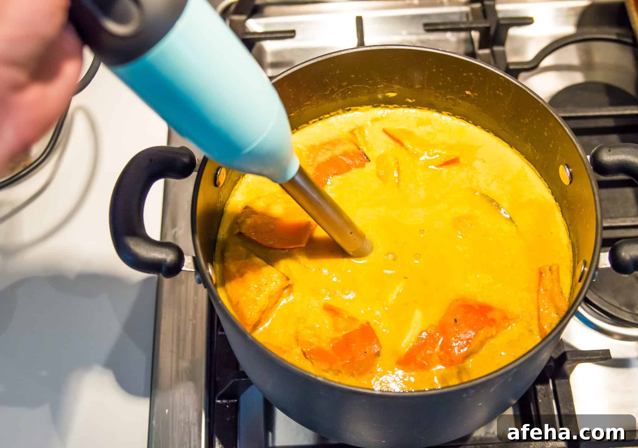 An orange soup in a pot on a stovetop with an immersion blender in it.