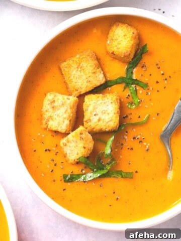 A bowl of butternut squash and red pepper soup garnished with croutons and fresh herbs.
