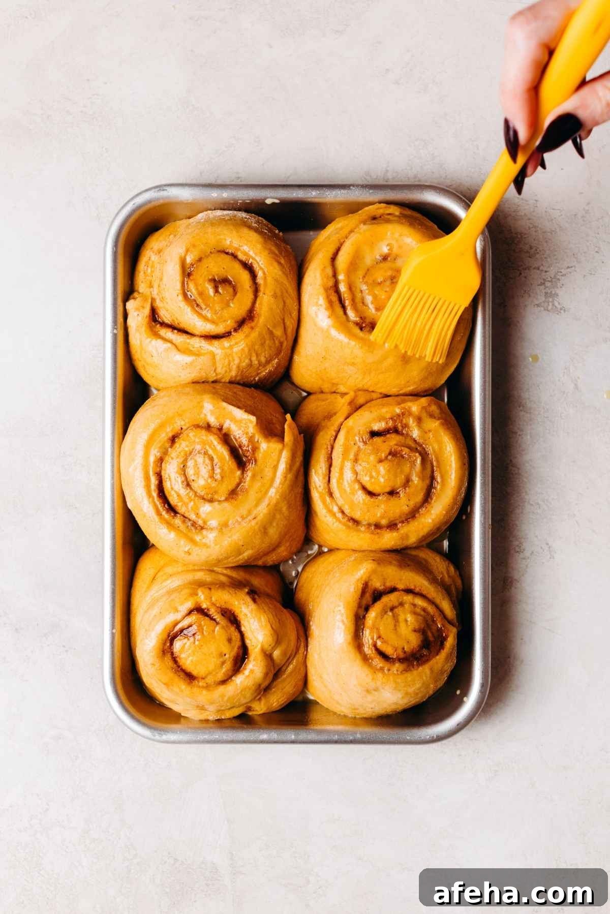 Brushing egg wash onto six pumpkin cinnamon roll slices on a metal baking sheet before baking.