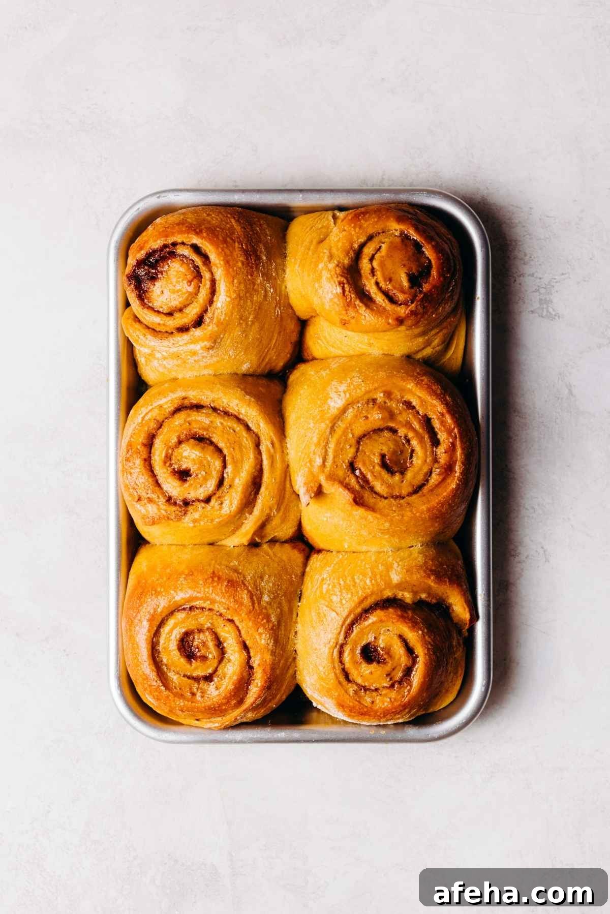 Six baked pumpkin cinnamon rolls on a metal baking sheet placed on a white and grey marbled background.