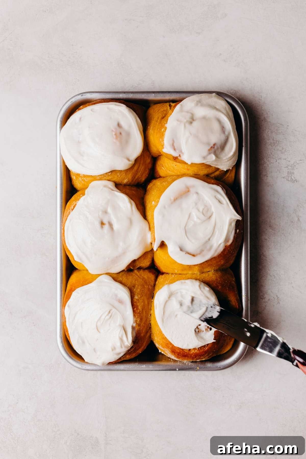 Pumpkin cinnamon rolls on a metal baking sheet being topped with maple frosting using an offset spatula.