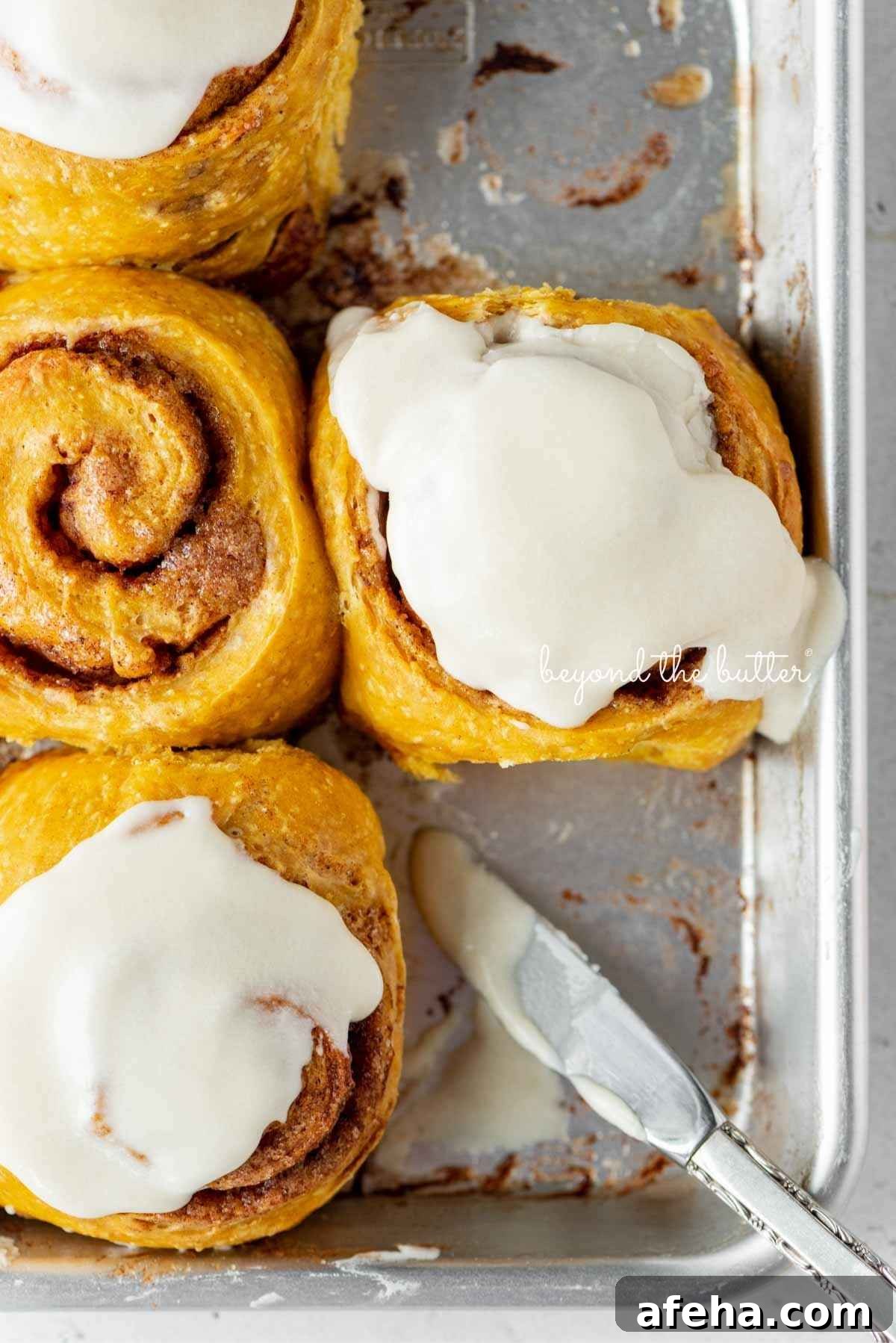 Four pumpkin cinnamon rolls on a small baking sheet with three topped with maple frosting using a small knife placed on a corner of the baking sheet.