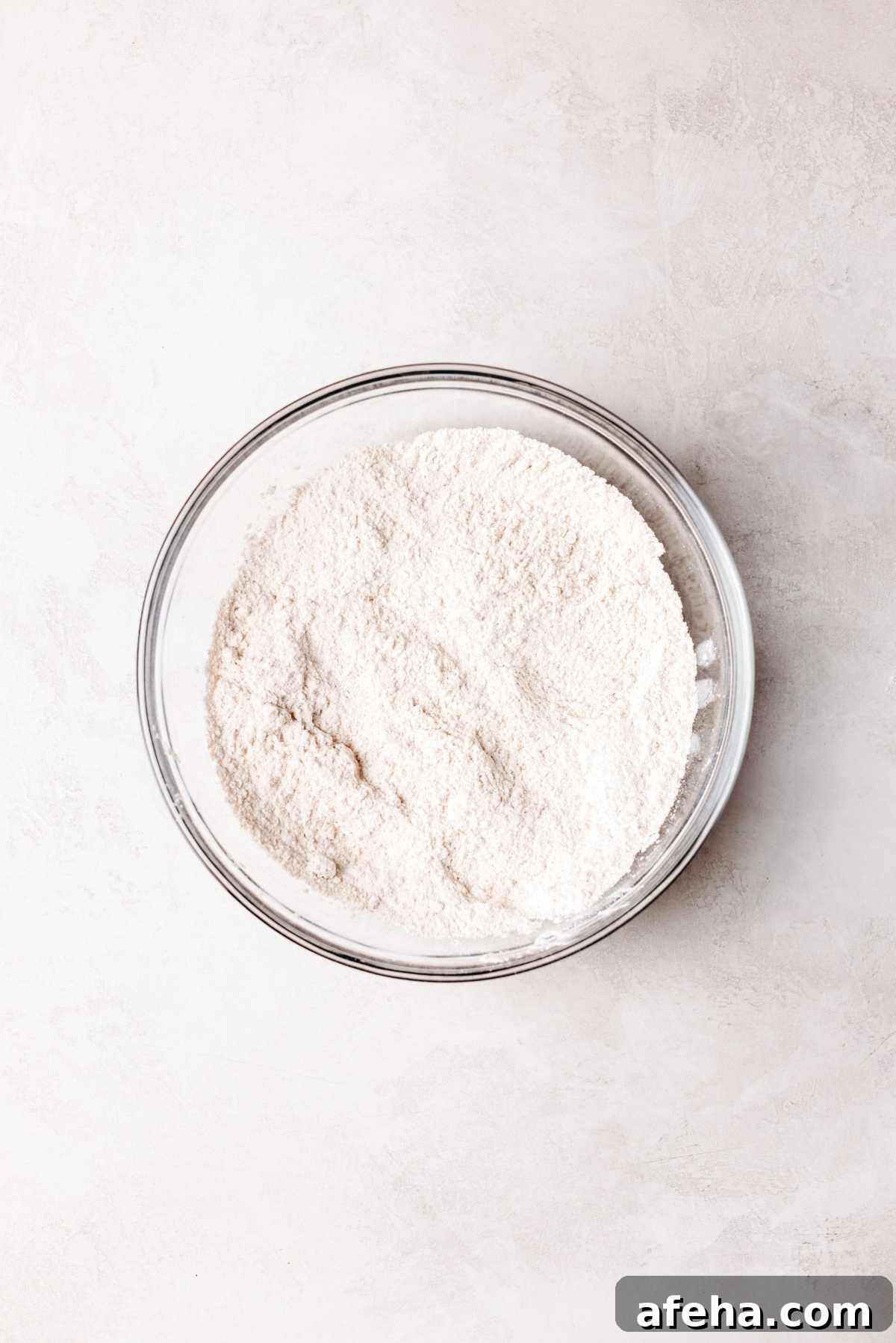 Whisked flour, yeast, brown sugar, cinnamon, and salt in a small glass mixing bowl on a white and grey marbled background.