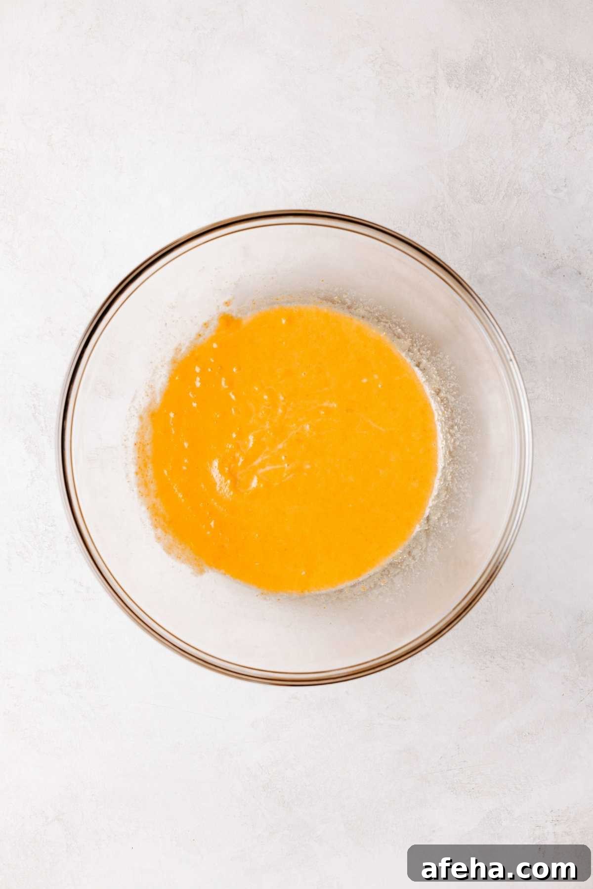 Whisked milk, pumpkin puree, melted butter, and egg in a medium size glass mixing bowl on a white and grey marbled background.