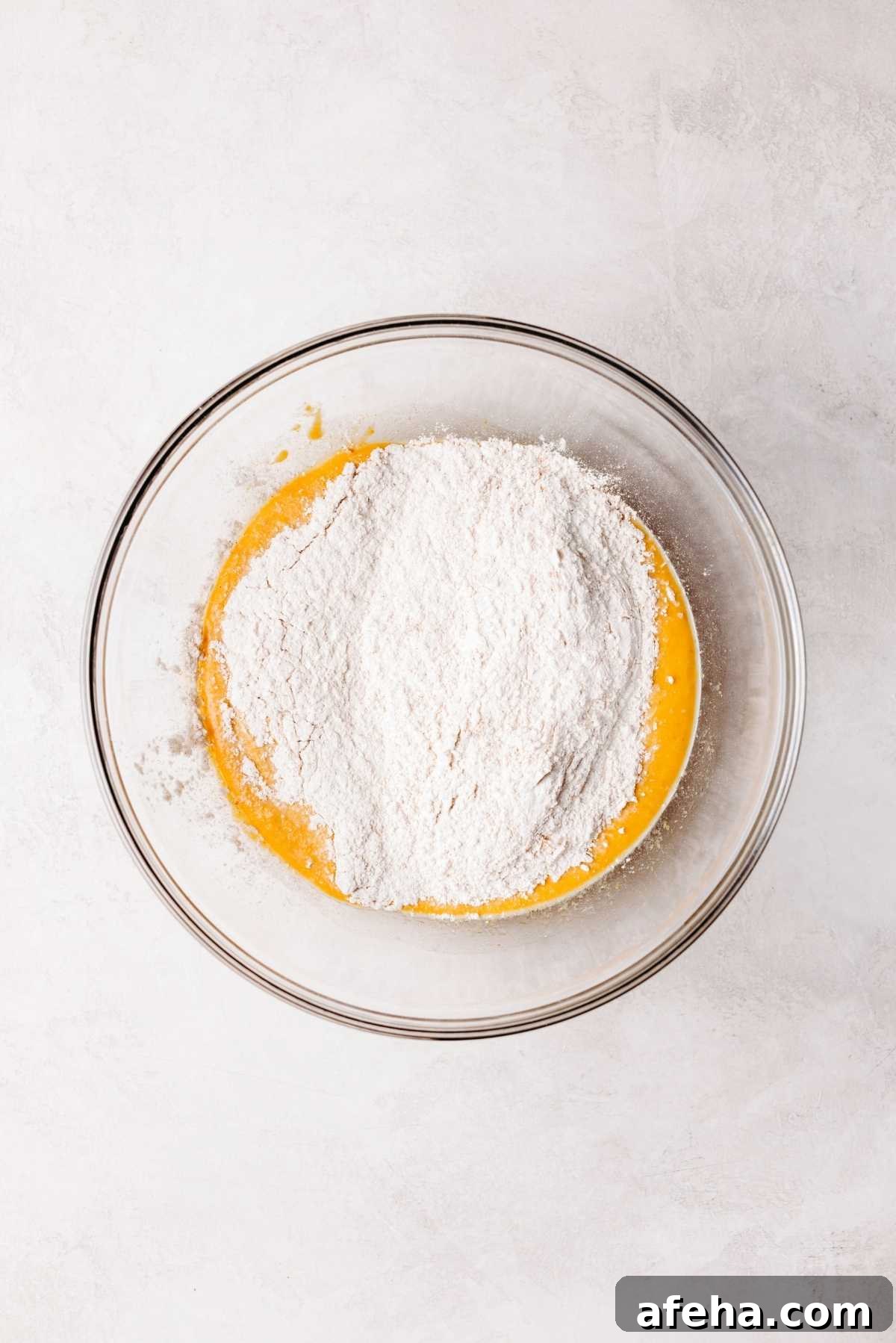 Flour mixture added to pumpkin cinnamon roll wet ingredients in a medium size glass mixing bowl on a white and grey marbled background.