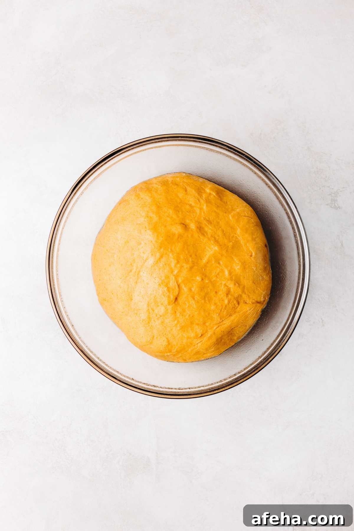 Pumpkin cinnamon roll dough doubled in size in a medium size glass mixing bowl on a white and grey marbled background.
