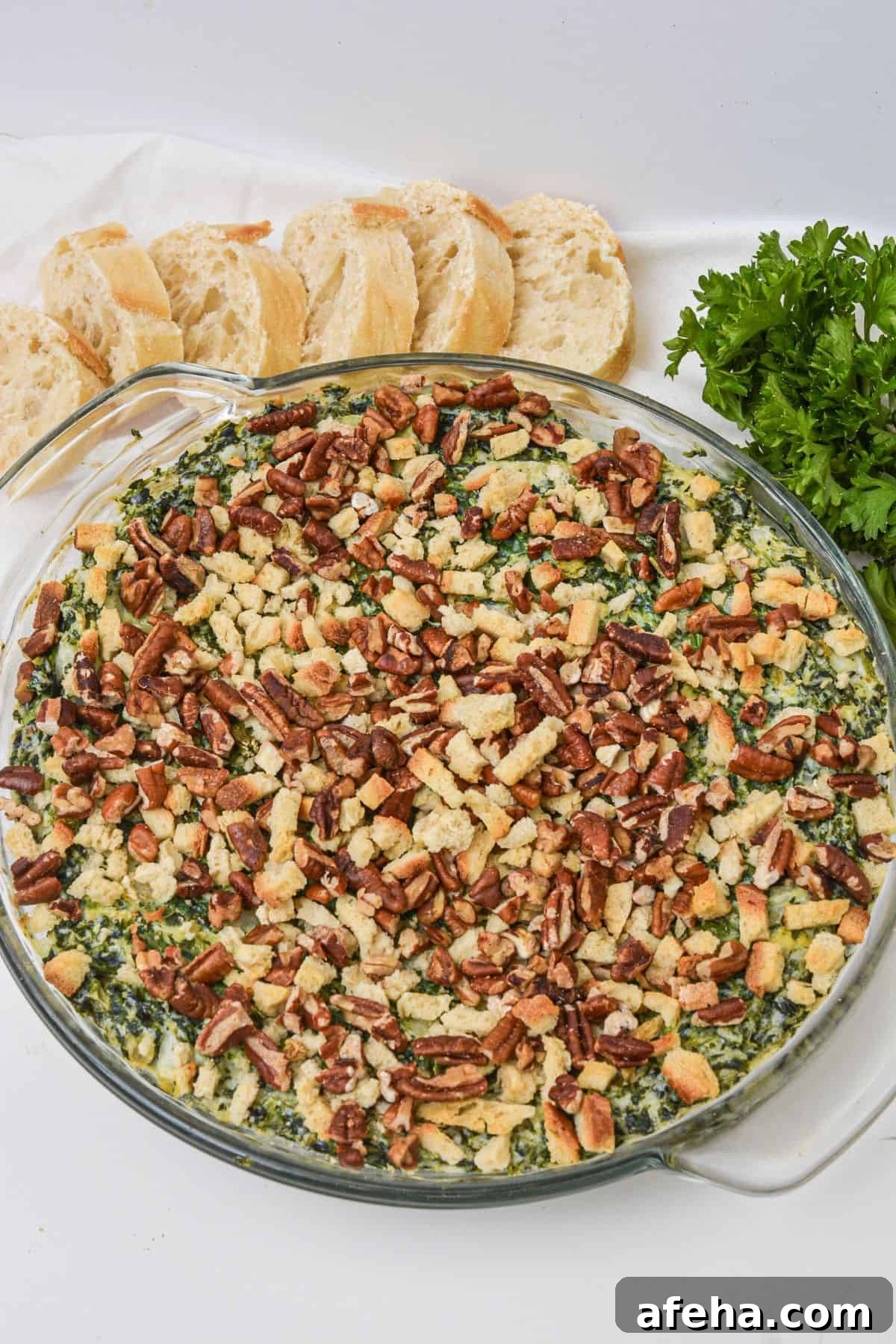 A round glass dish filled with artichoke and spinach dip recipe, topped with chopped pecans and breadcrumbs, placed next to sliced bread and fresh parsley.