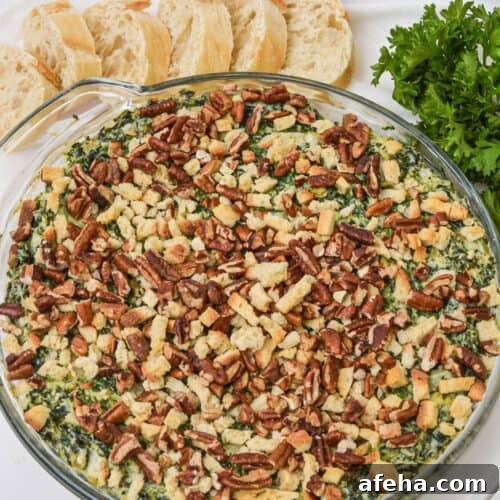 A round glass dish filled with a baked Artichoke and Spinach Dip Recipe, topped with chopped pecans and breadcrumbs, served alongside sliced baguette and fresh parsley.
