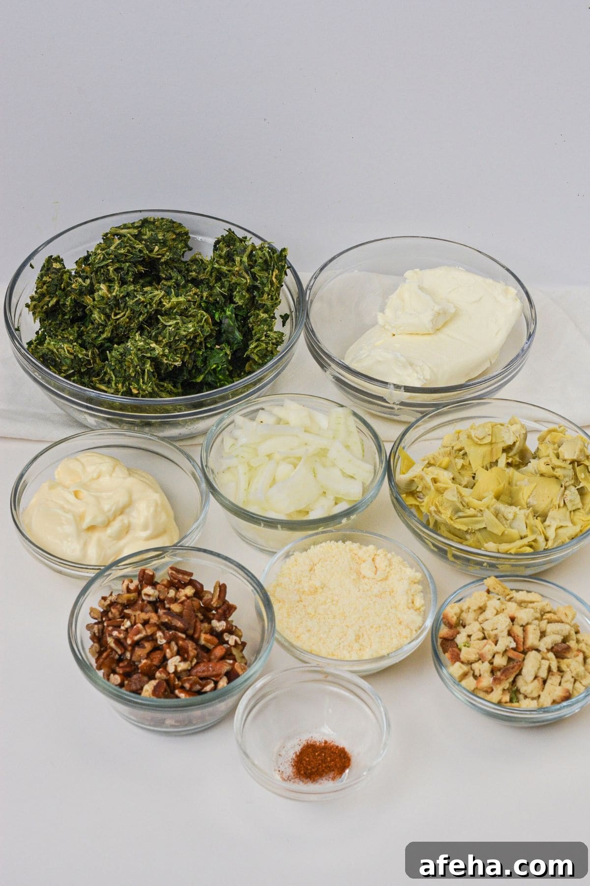 Eight glass bowls filled with chopped kale, cream cheese, mayonnaise, sliced onions, artichokes, pecans, grated cheese, croutons, and spices.