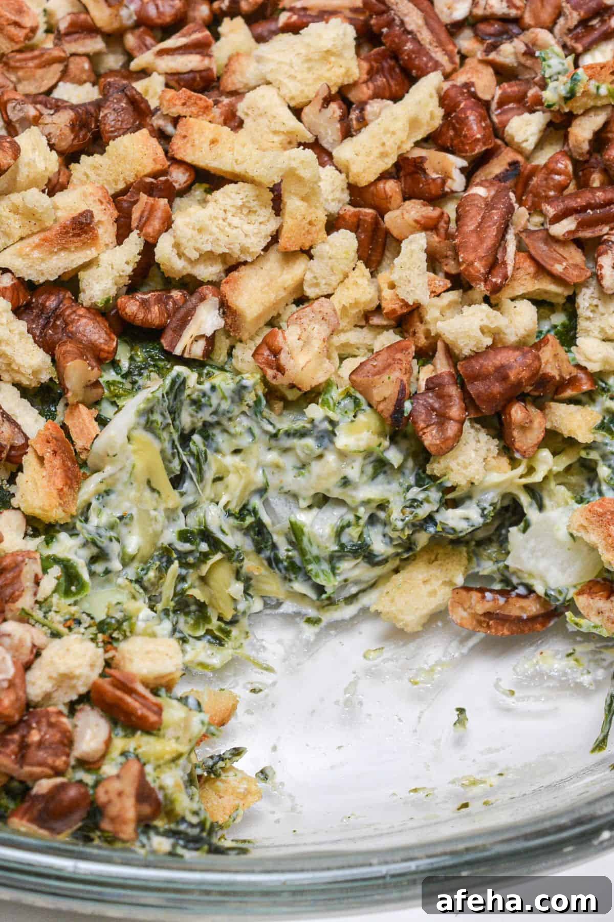 A close-up of a spinach casserole inspired by an Artichoke and Spinach Dip Recipe, topped with chopped pecans and croutons, with a portion removed to reveal the creamy spinach filling.