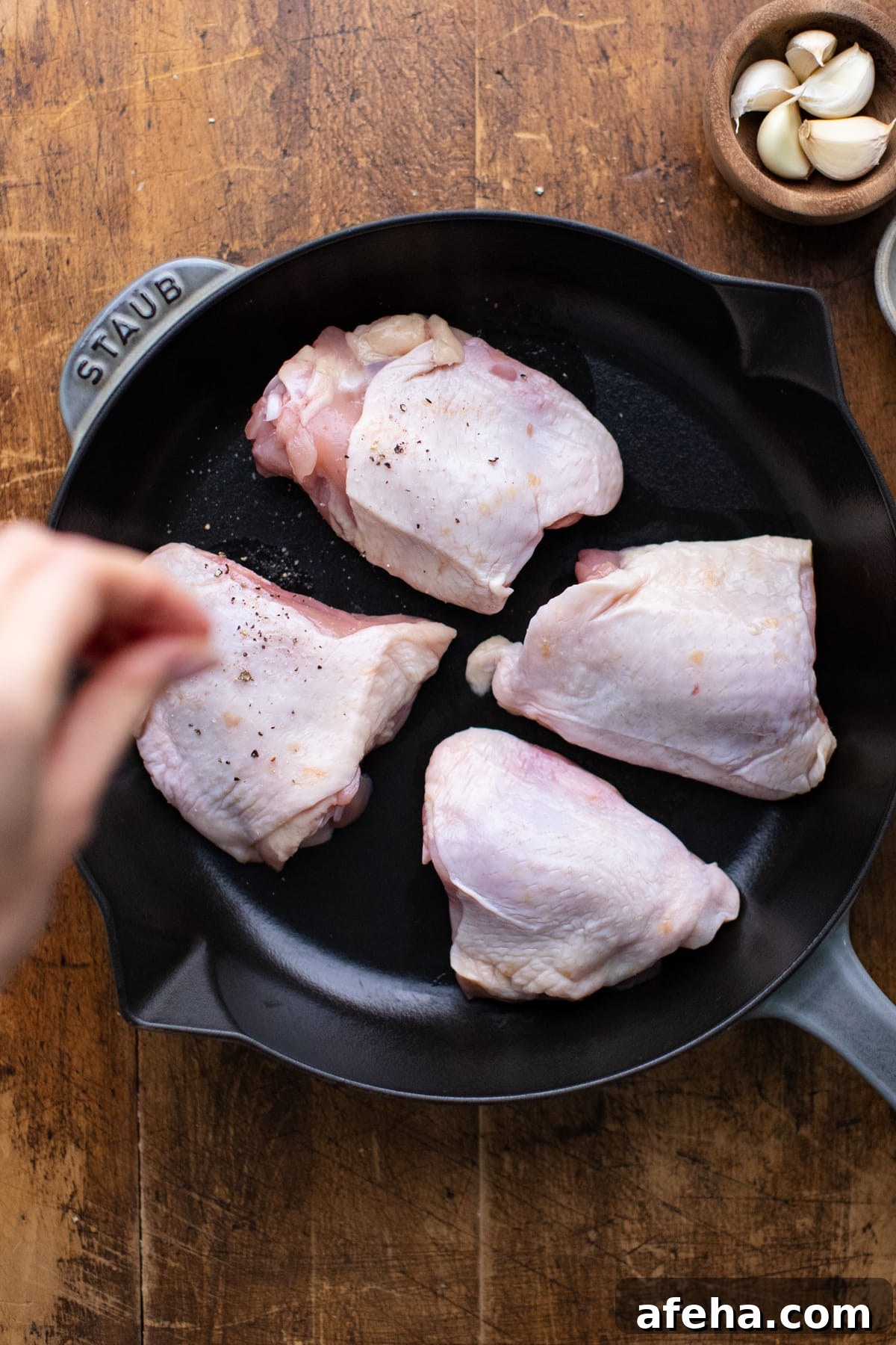 Hand sprinkling salt and pepper over chicken thighs in a cast iron skillet sitting on wooden backrgound.