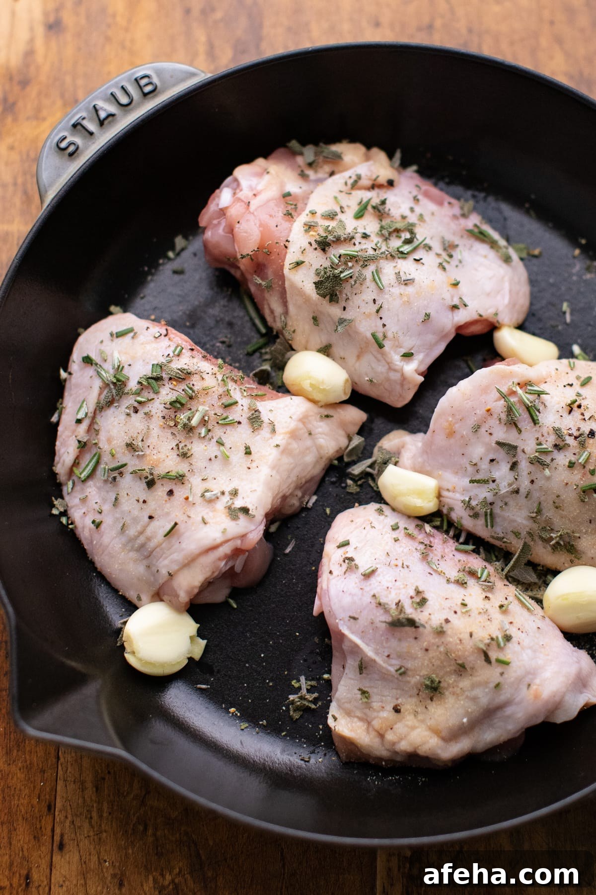 raw chicken thighs covered with chopped herbs and garlic cloves in a cast iron pan on a wooden background.