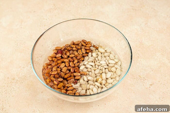 A glass bowl features uncooked tepary beans on the right, alongside white beans, all resting on a light surface.