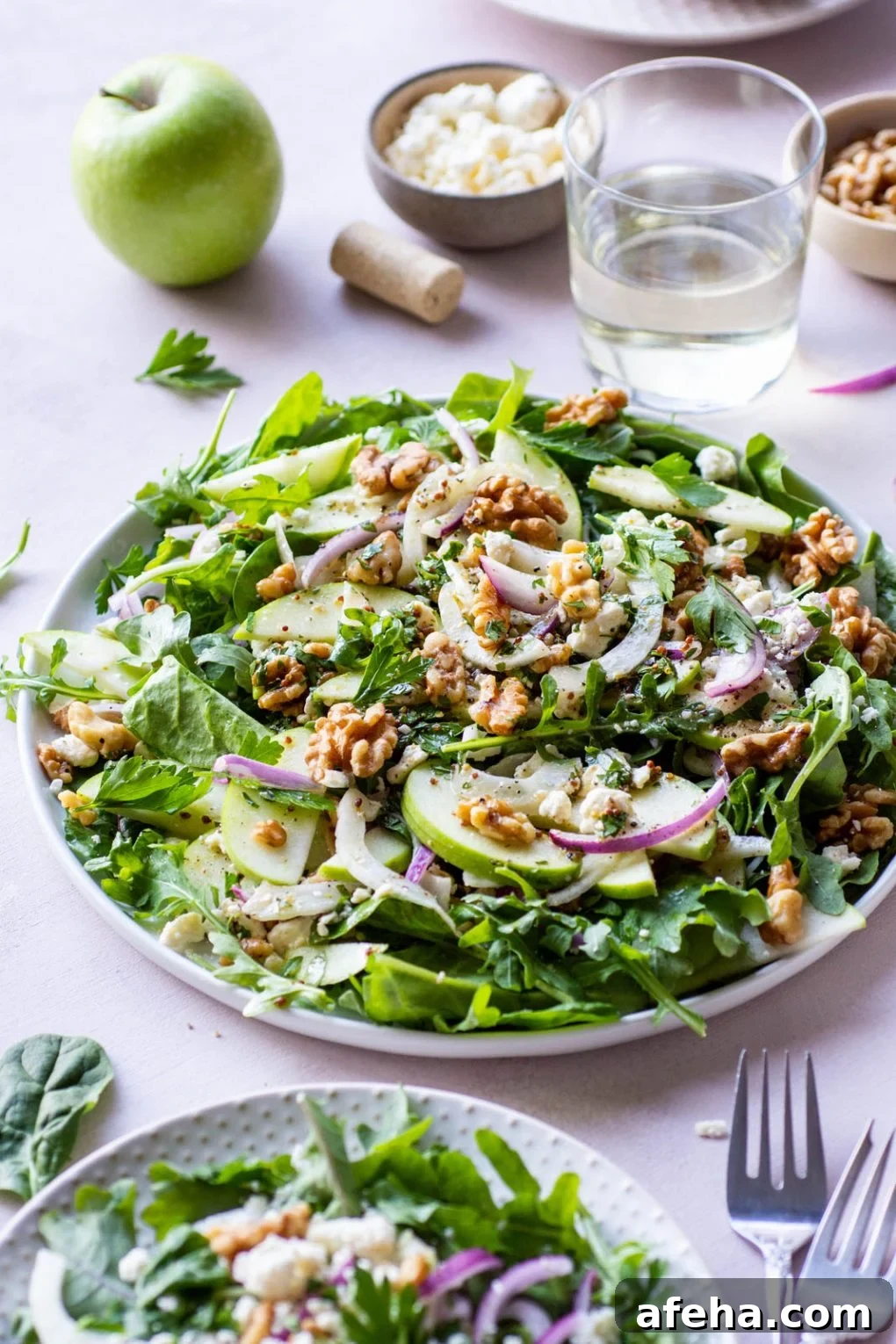 Side angle shot of a large green salad on a round plate and a light colored background. Salad greens layered with thinly sliced green onion, thinly shaved fresh fennel, sliced red onion, walnuts, and torn parsley leaves. Drizzled with a mustardy dressing. Next to a smaller plate of salad with a fork on the plate. 