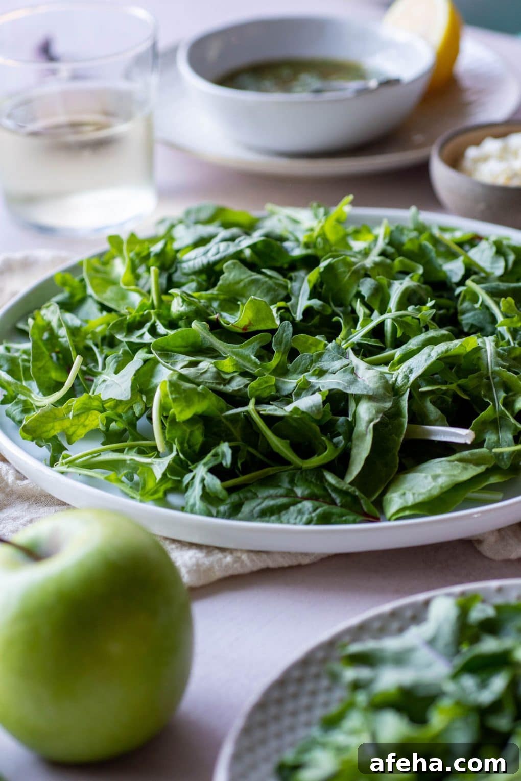 Side angle shot of a plate of salad greens on a light colored background with a green apple in the foreground of the image.