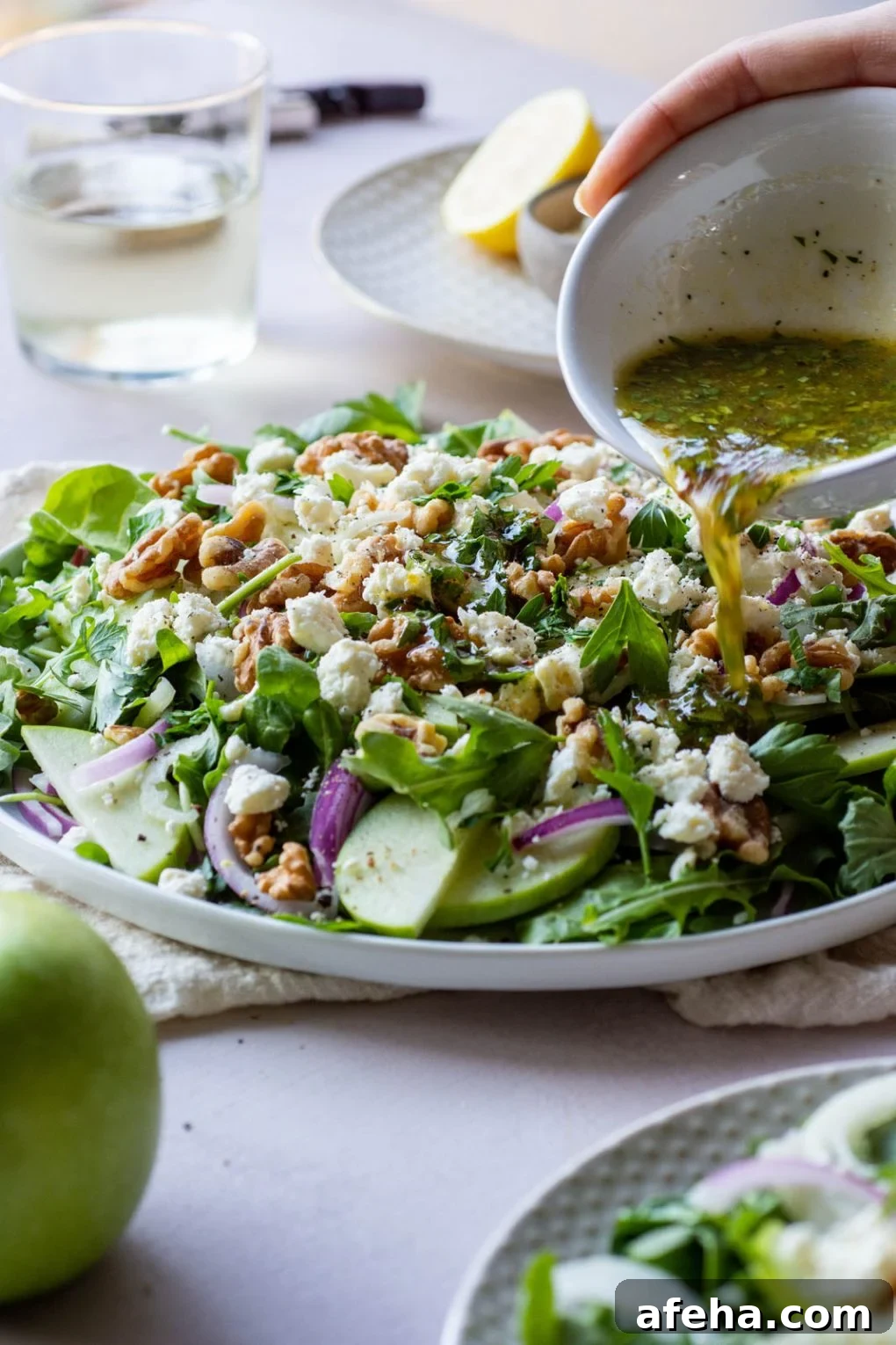 Side angle shot of dressing being drizzled over layered salad of greens, apples, fennel, walnuts, red onion, and parsley.