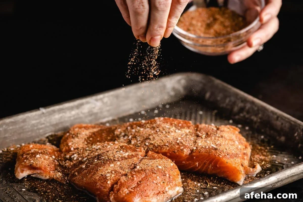Salmon fillets on a baking sheet with a hand sprinkling a spice blend onto them from a small bowl.