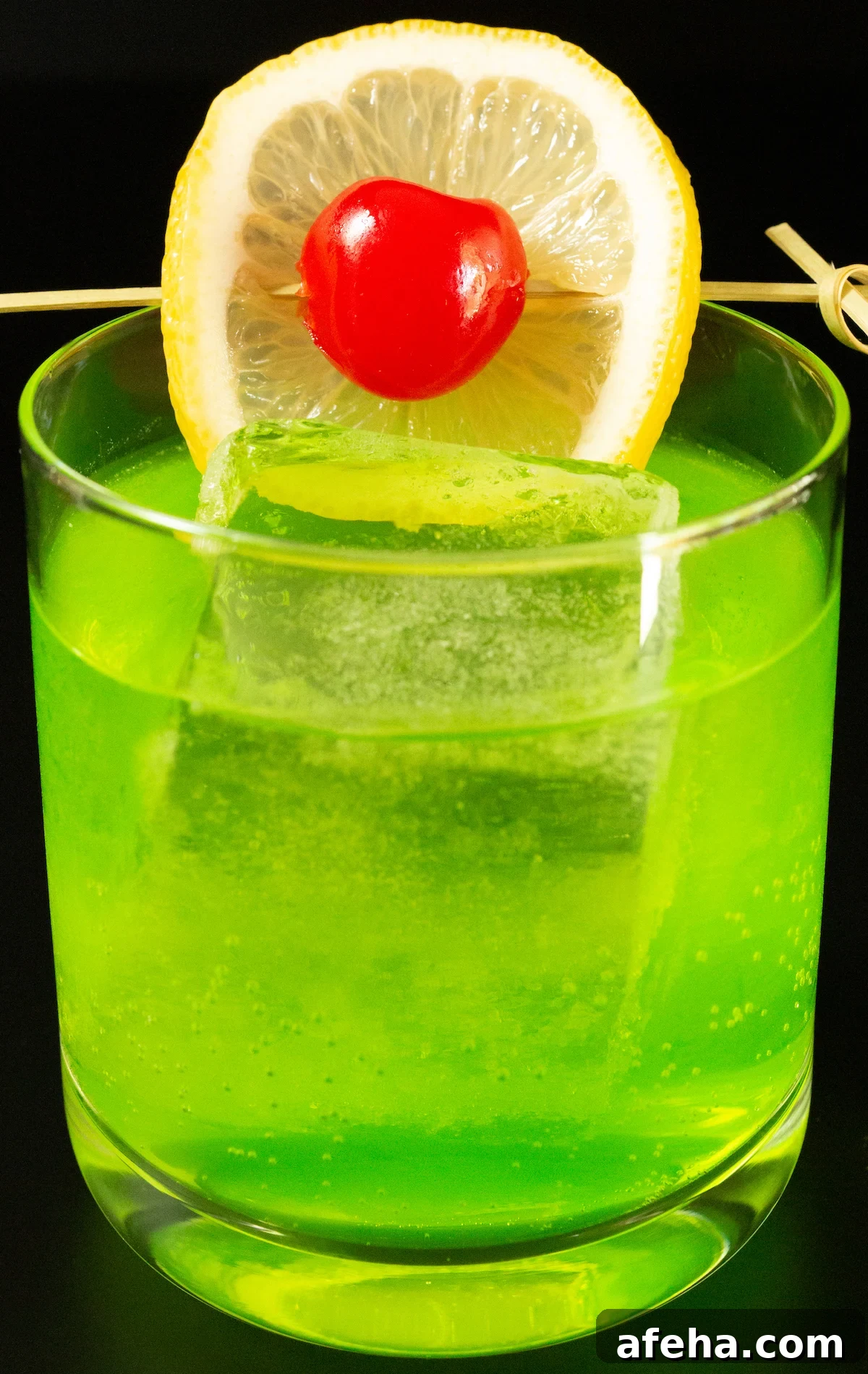 A lowball glass with a large block of ice is filled with a bright neon green Midori Sour cocktail that's garnished with a lime wheel and a maraschino cherry on a skewer.
