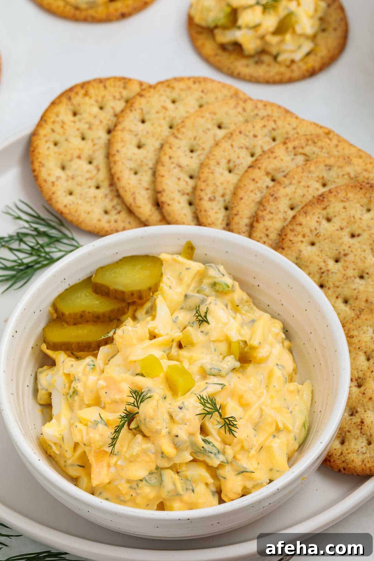 Bowl of dill pickle egg salad garnished with sliced pickles and fresh dill, served with round crackers arranged on a plate for snacking.