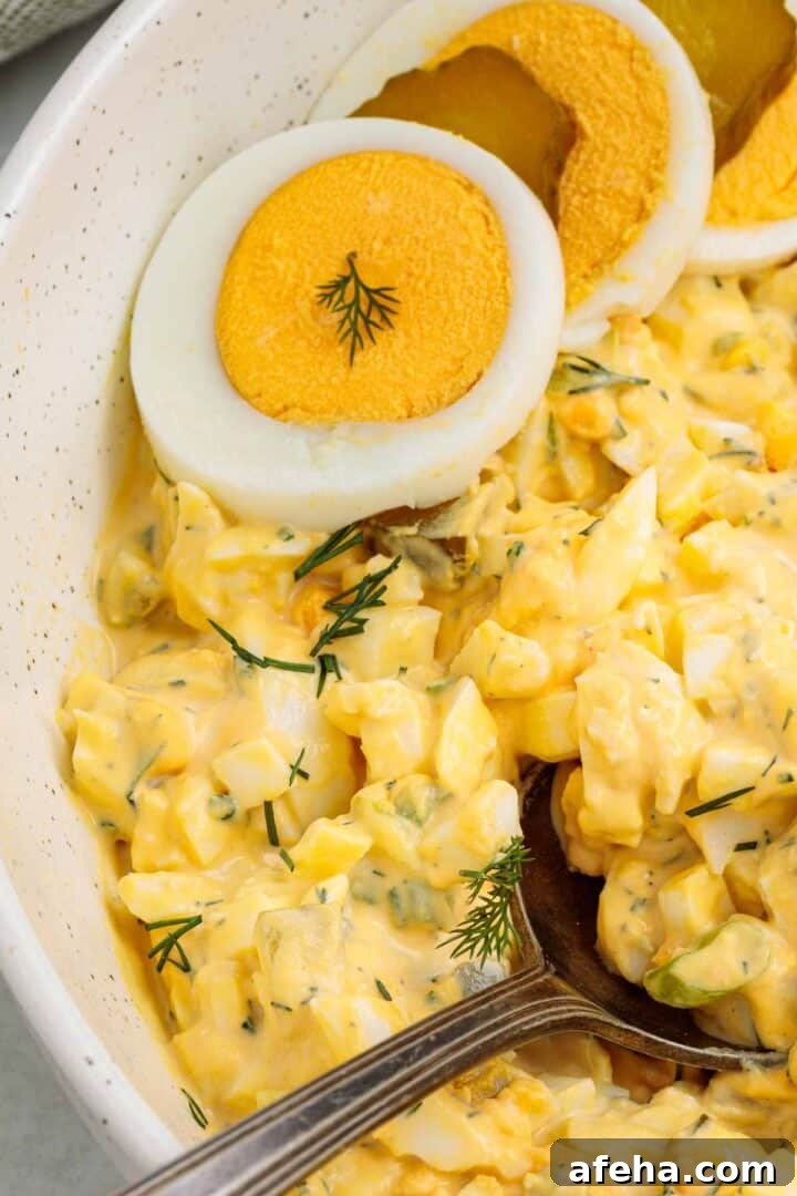 Close up of creamy dill pickle egg salad with chopped eggs, pickles, and fresh dill, topped with sliced hard boiled eggs and served in a white bowl with a spoon.