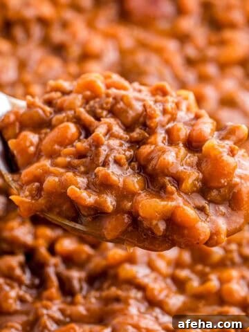 Spoonful of baked beans with ground beef held above the dish to show the thick texture and rich sauce. Tender beans and crumbled beef are visible in the close up.