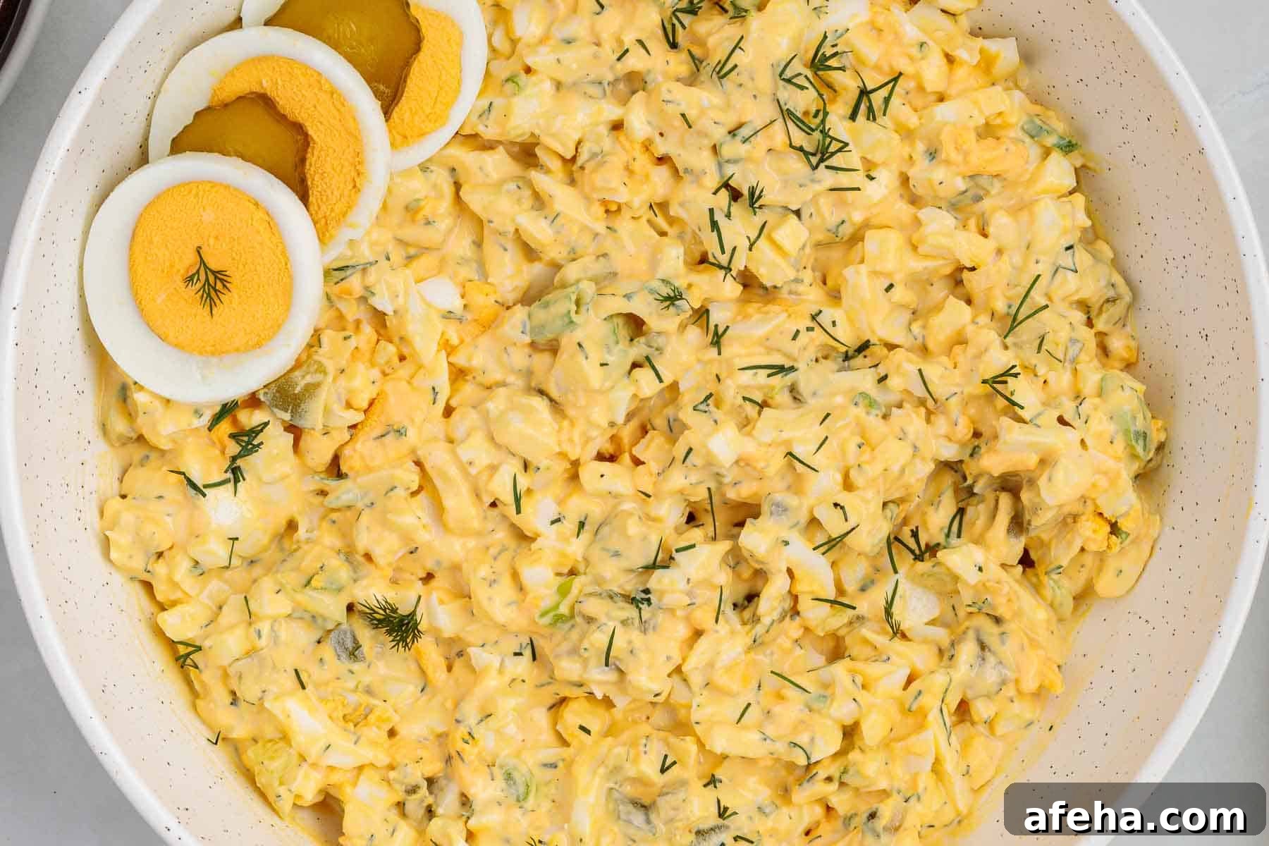 Overhead view of a large bowl filled with dill pickle egg salad mixed with chopped eggs, pickles, and fresh dill, with sliced hard boiled eggs arranged on top.