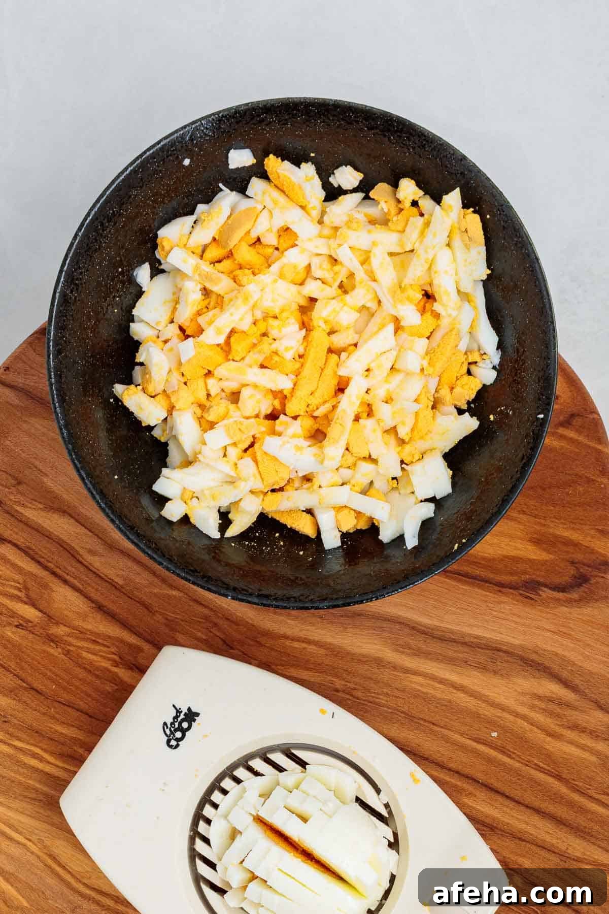 Chopped hard boiled eggs in a bowl with visible yolk and whites, with an egg slicer on a wooden cutting board showing part of the prep process.