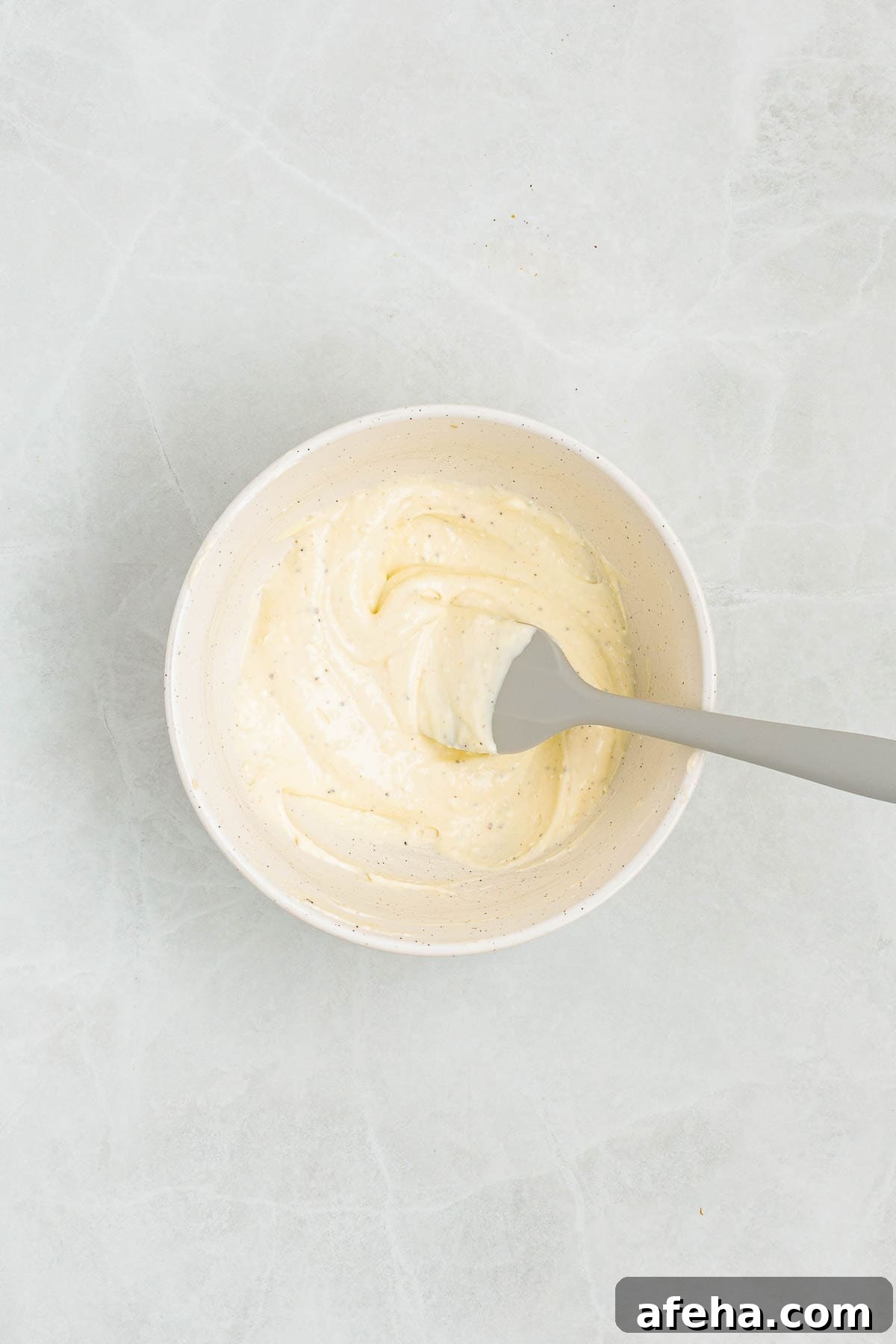 Mixing bowl of smooth creamy dressing made with mayonnaise, whipped cream cheese, dijon, and seasonings, being mixed with a spatula on a light countertop.