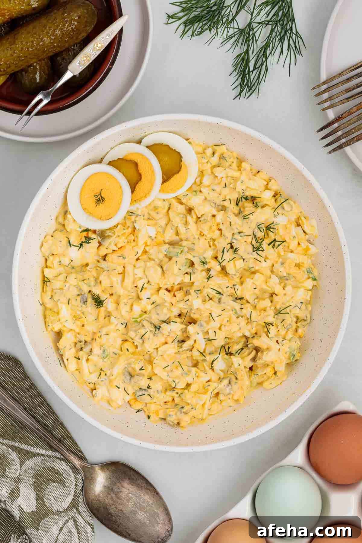 Large serving bowl of dill pickle egg salad garnished with sliced hard boiled eggs and fresh dill, surrounded by pickles, utensils, and whole eggs on a table.