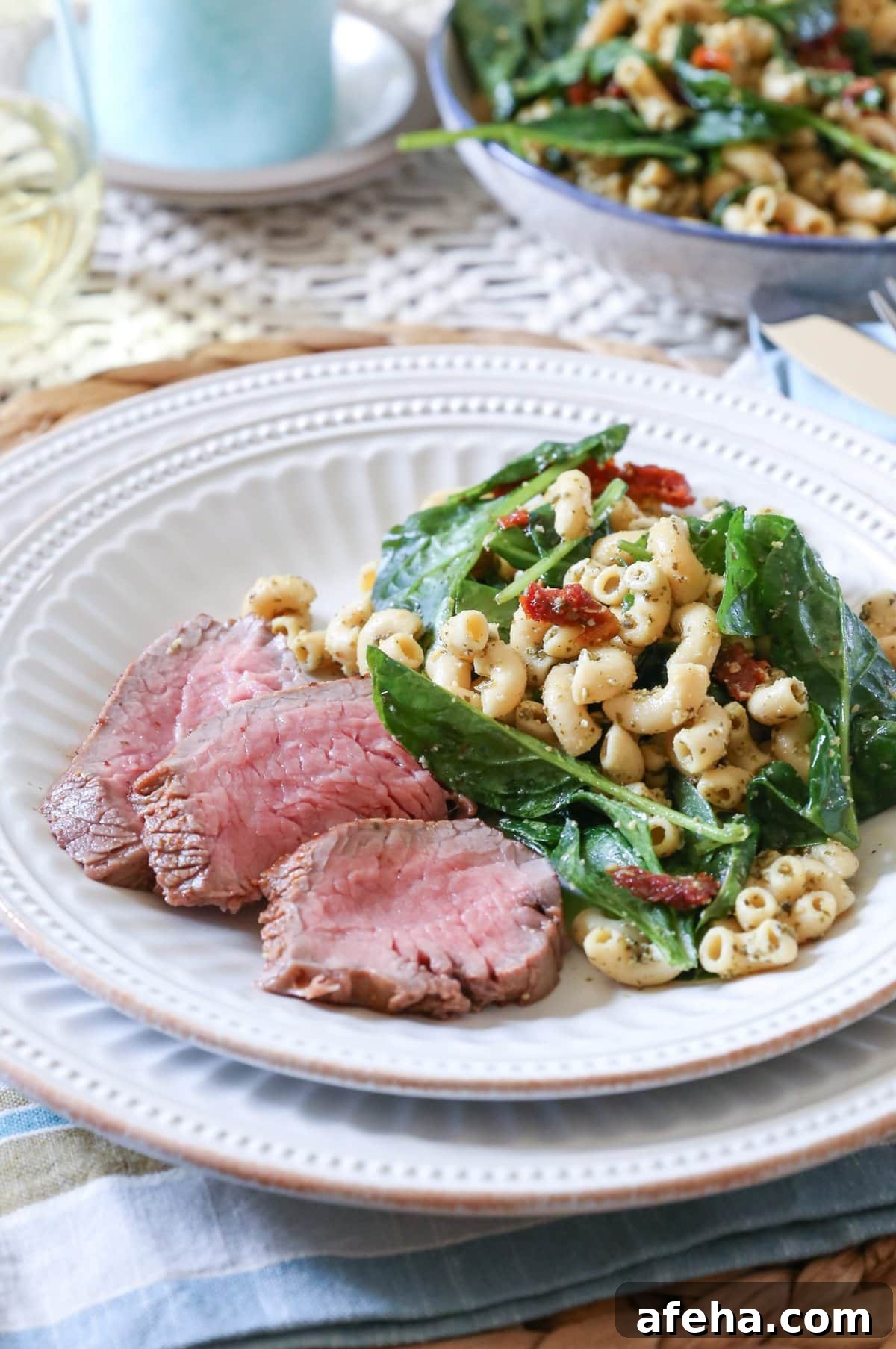Slices of oven baked tri tip on a white plate with macaroni salad. A place mat and napkin underneath the plate and a glass of wine in the background.