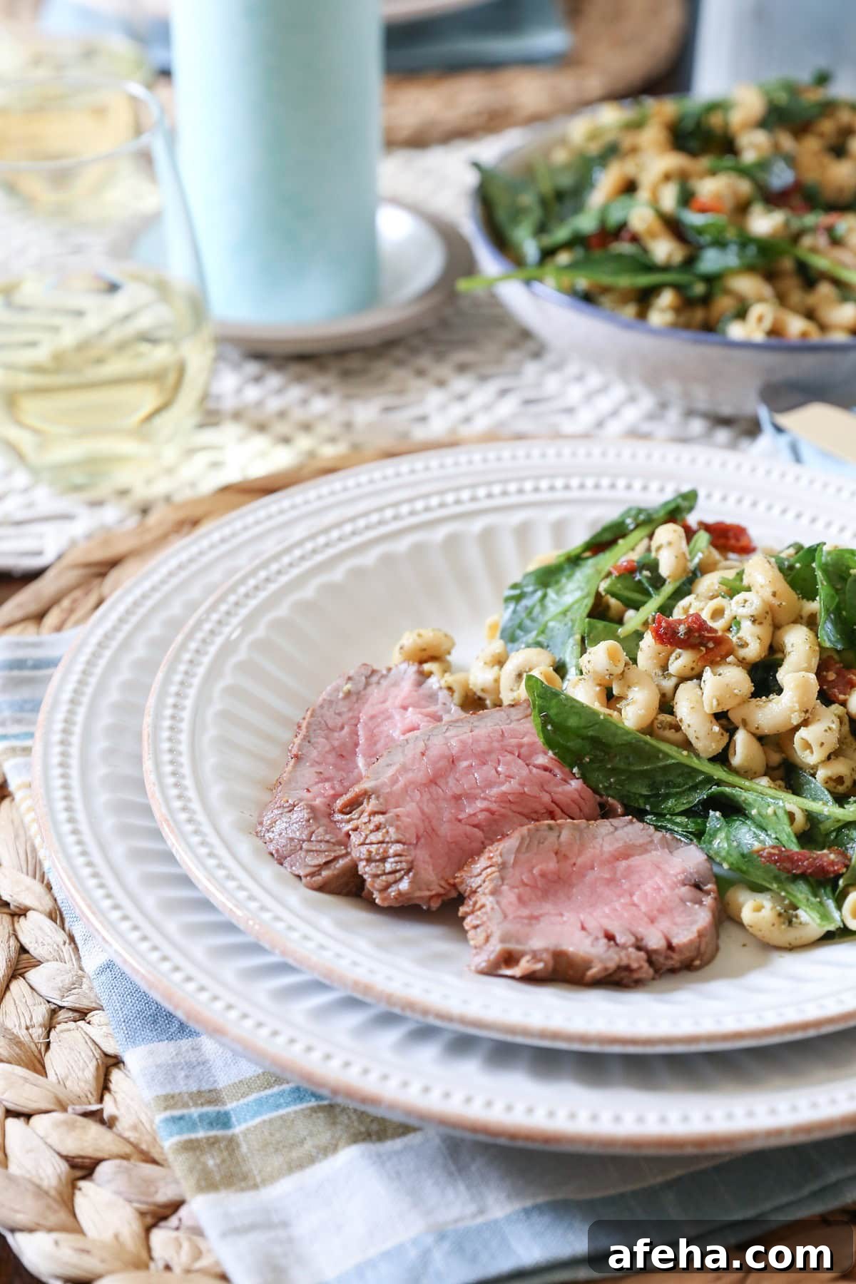 Tri tip slices on a white plate next to macaroni salad, ready to serve. Plate is on top of a placemat with a napkin and there's a bowl of macaroni salad in the background.