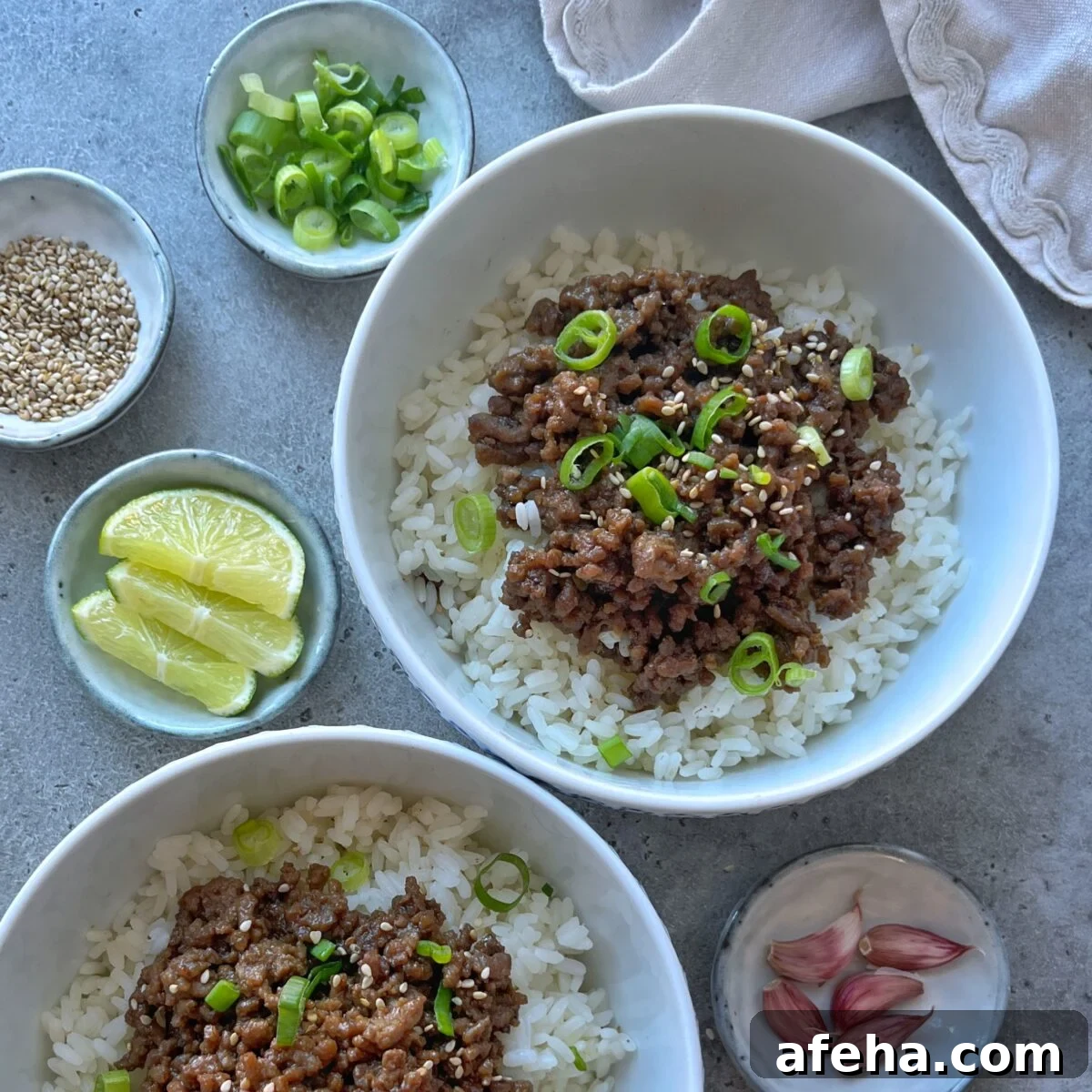 Mongolian Beef with Ground Beef: Quick Savory Stir-Fry Recipe 3 Two bowls of white rice topped with savory Mongolian Ground Beef and chopped green onions, surrounded by small dishes of lime wedges, sesame seeds, green onions, and garlic cloves.