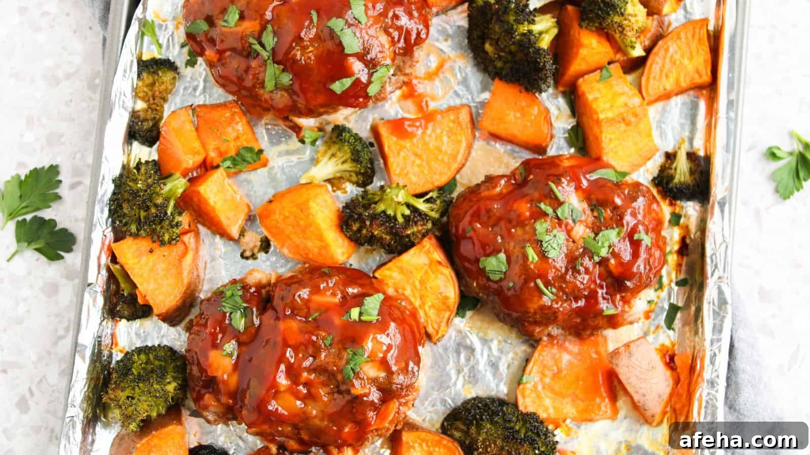 Three mini meatloafs topped with sauce are baked on a foil-lined tray with roasted sweet potato chunks and broccoli florets, garnished with chopped parsley.