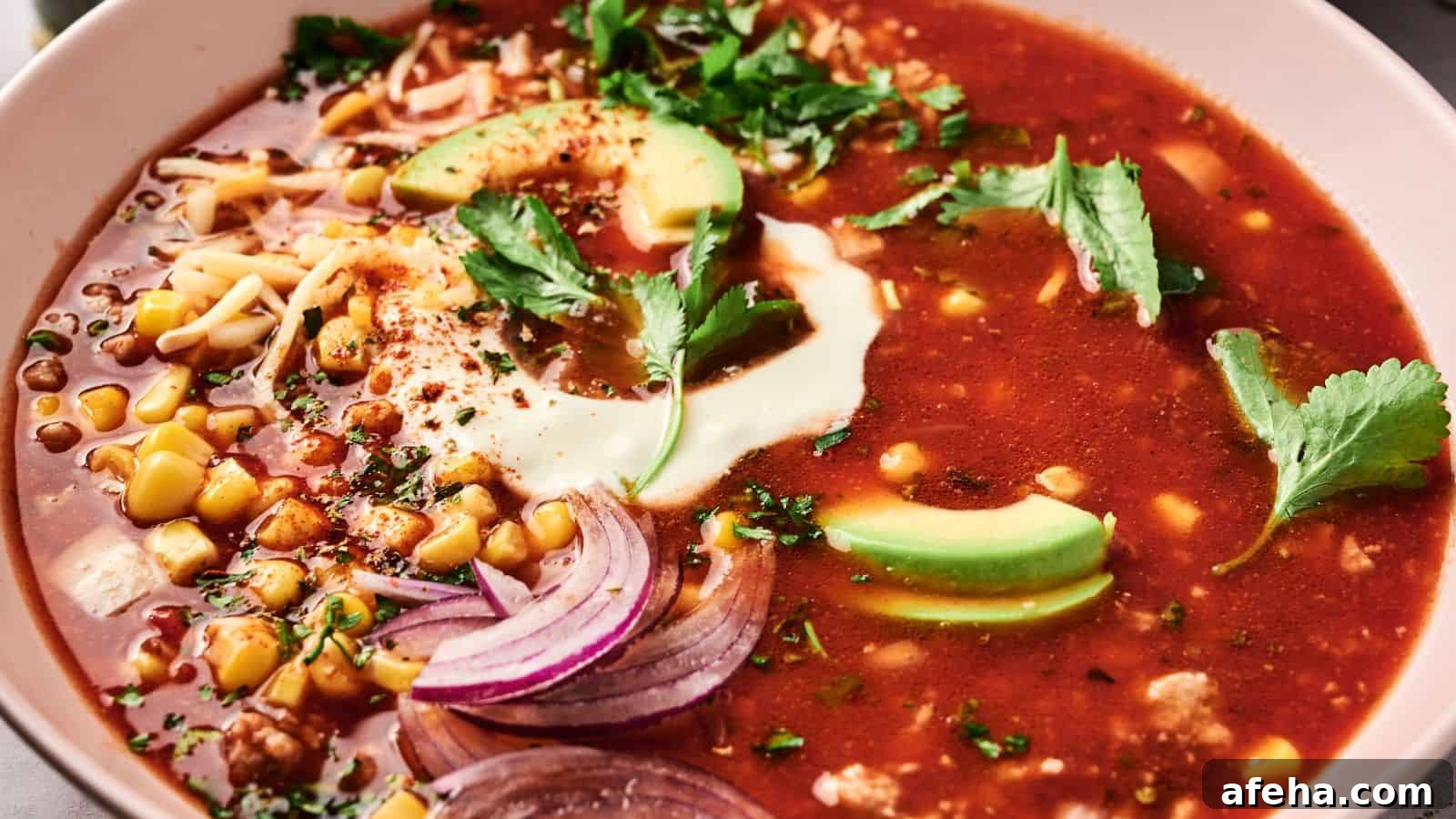 A bowl of red soup topped with avocado slices, sour cream, cilantro, sliced onions, corn, and shredded cheese.
