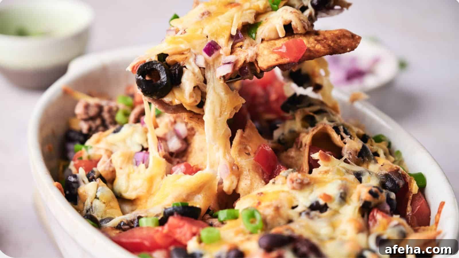 A bowl of taco casserole topped with melted cheese, black olives, tomatoes, and green onions, with a creamy dip in the background.