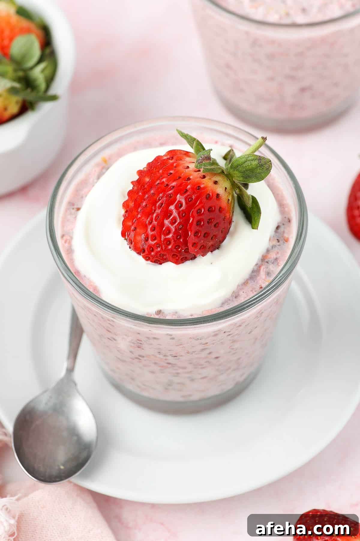 Glass jar of strawberry overnight oats, topped with yoghurt and a strawberry, sitting on a white saucer with a spoon on edge.
