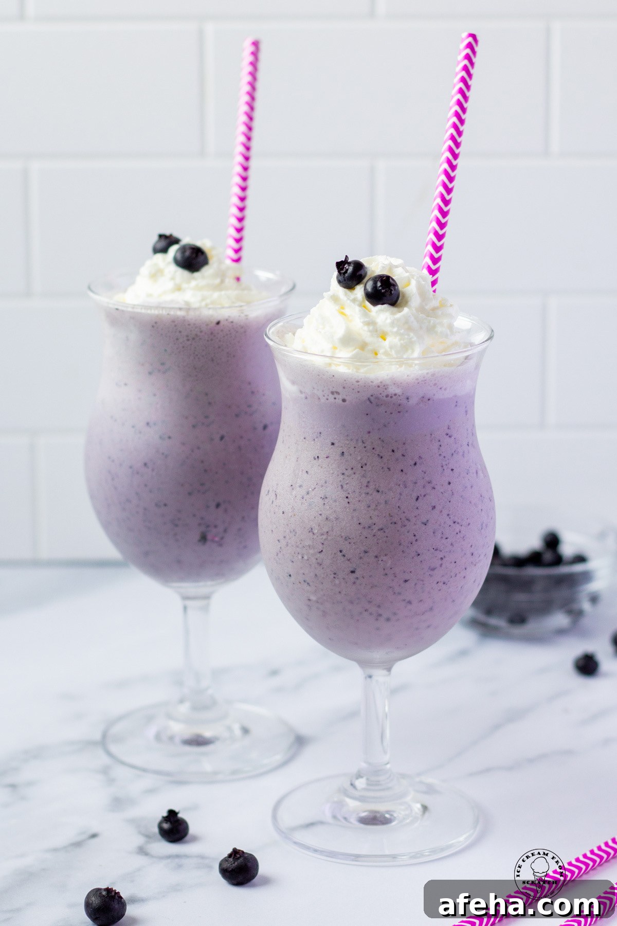 Two blueberry milkshakes in hurricane glasses with purple and white striped straws
