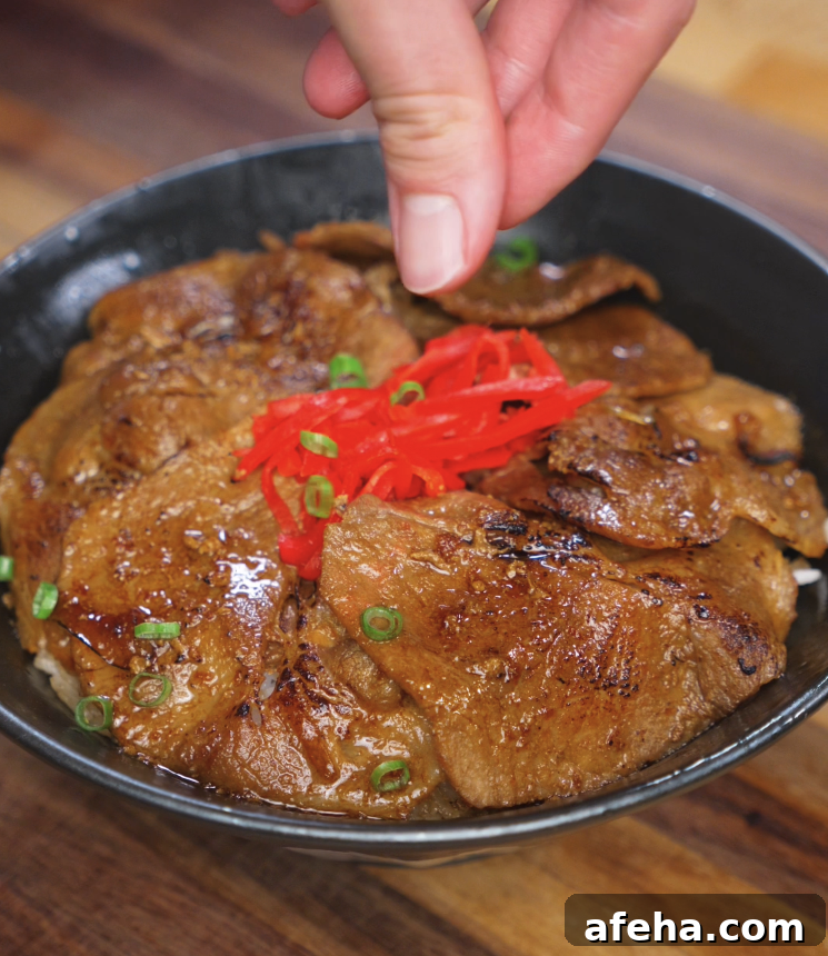 A hand sprinkles vibrant red pickled ginger and scallions over a bowl of glazed pork, finishing off the dish with color and freshness.