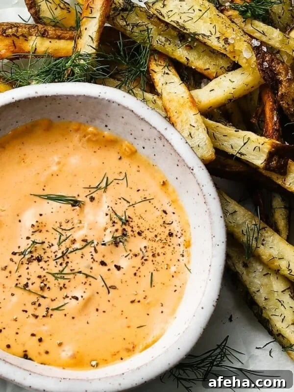 A bowl of chipotle mayo garnished with herbs beside crispy seasoned fries.