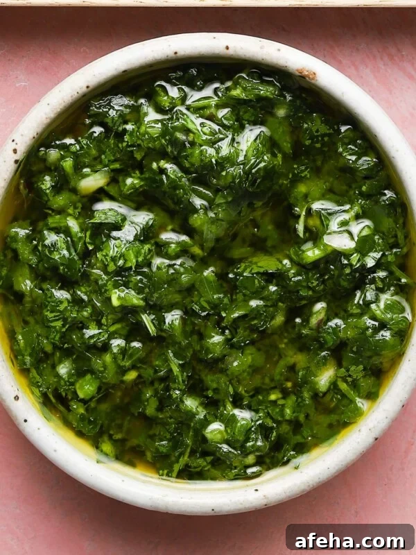 A bowl of vibrant green chimichurri sauce on a pink surface.