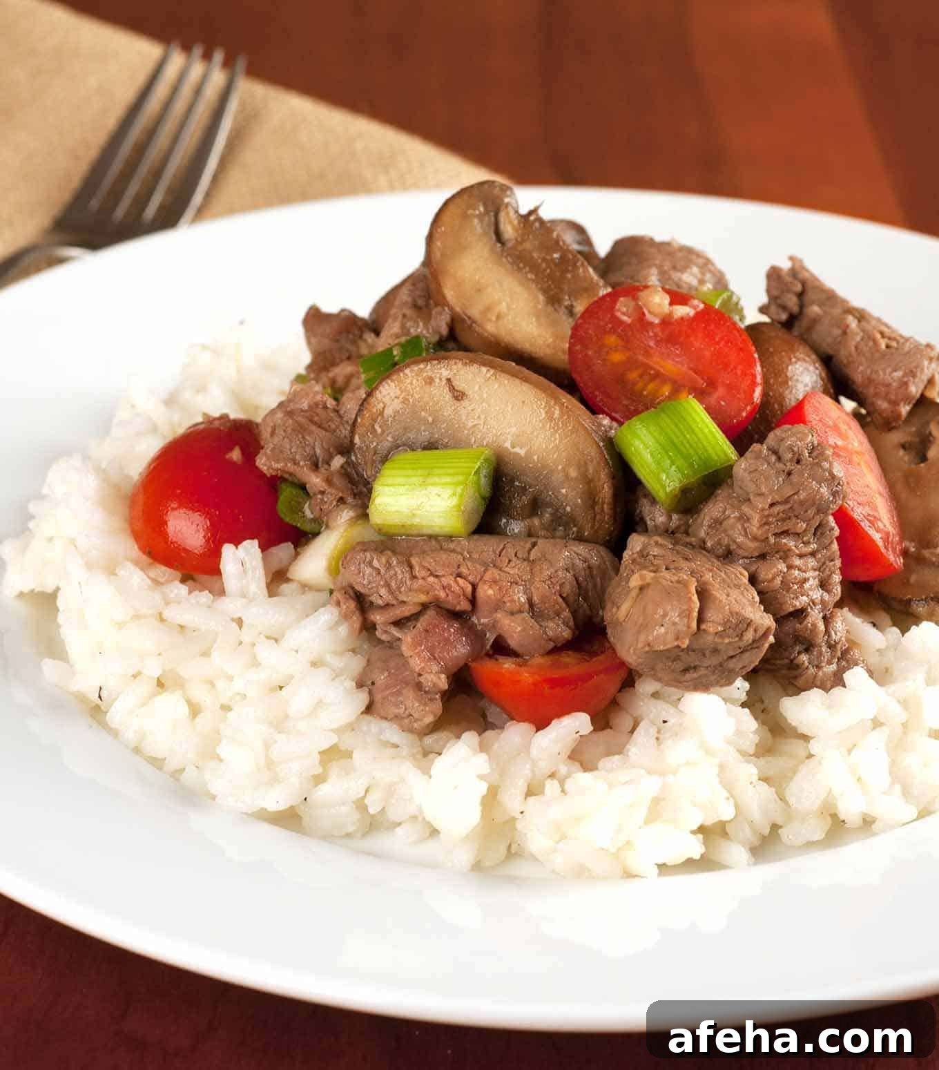 Tenderloin beef tips sautéed with mushrooms, cherry tomatoes, and scallions served over a bed of white rice on a white plate.