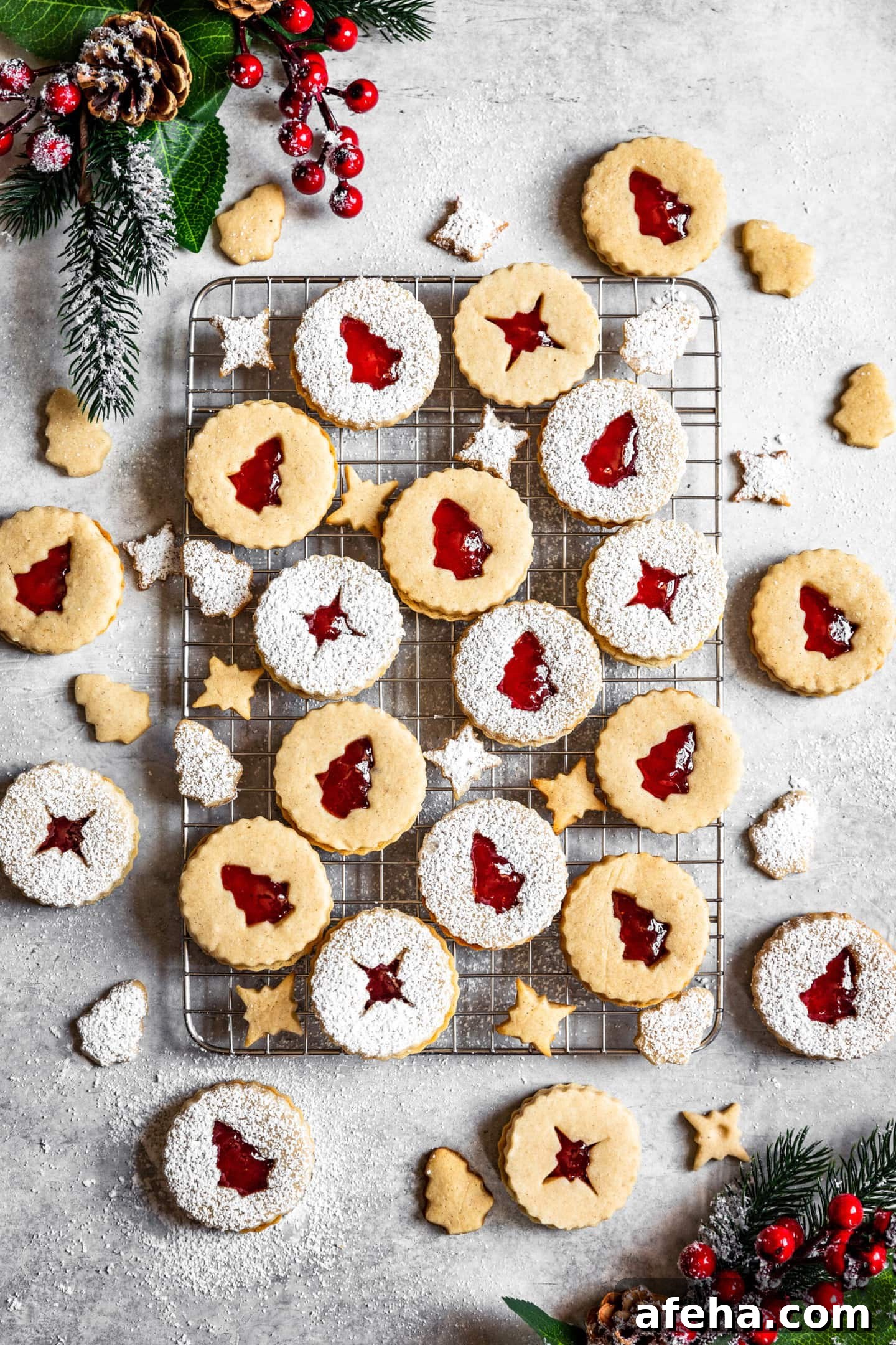 Strawberry Cardamom Linzer Cookies dusted with powdered sugar on top of a cooling rack surrounded by pine tree branches, holly berries, and pine cones.