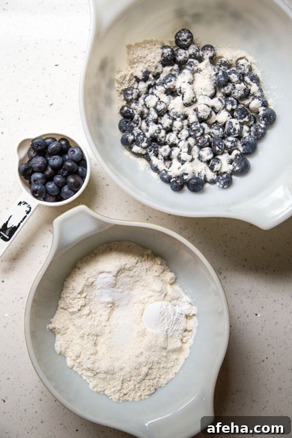 A mixing bowl with blueberries and flour next to another mixing bowl with a flour mixture next to a measuring cup with some fresh blueberries