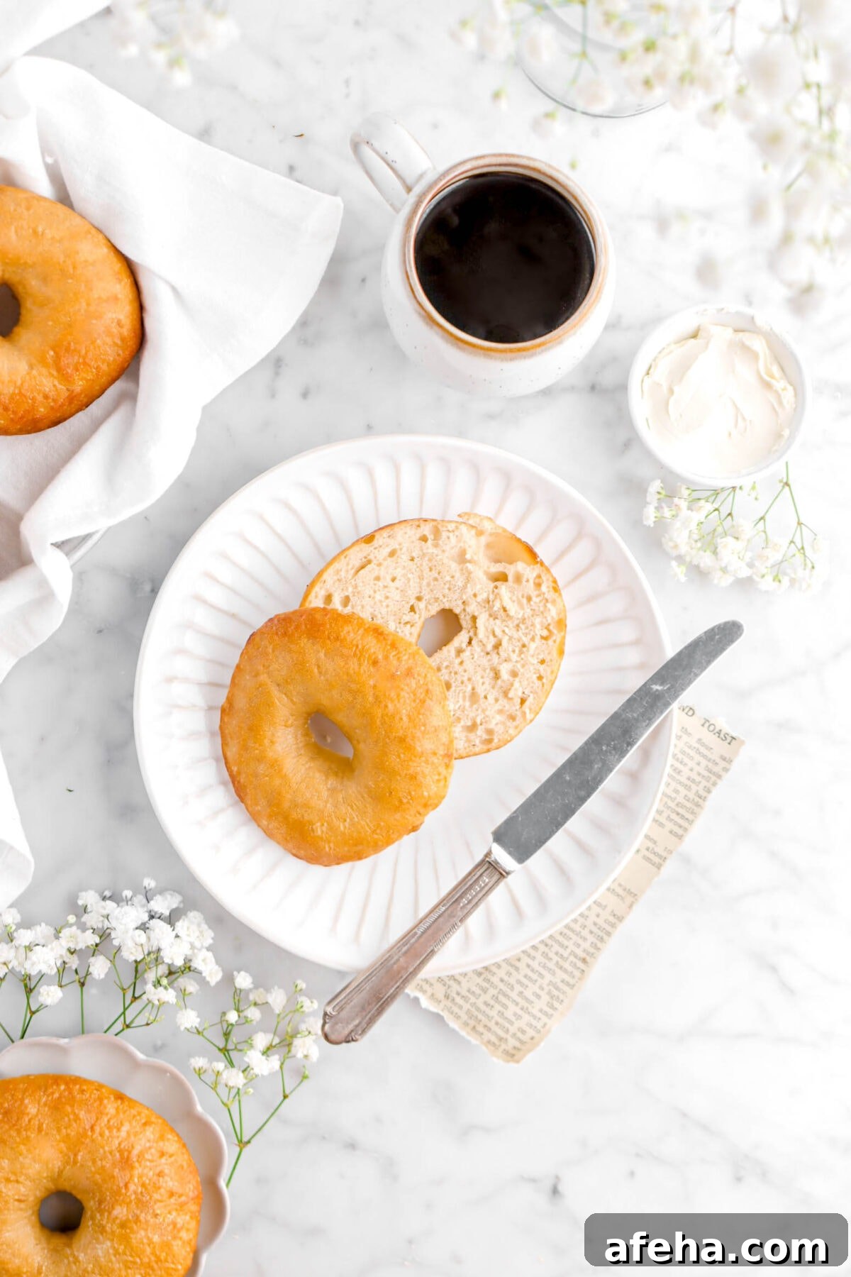 overhead shot of cut open bagel on white plate with knife, bowl of cream cheese, flowers, and cup of coffee