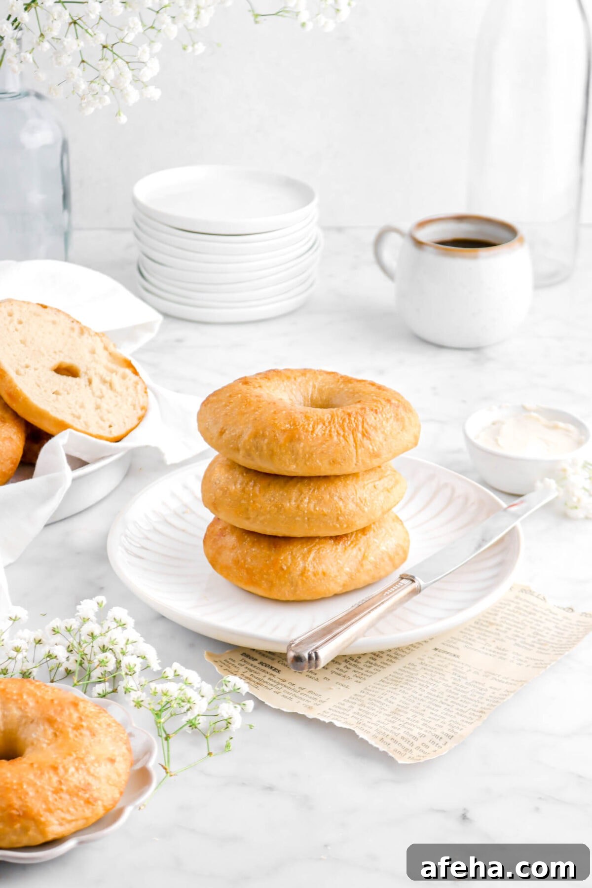three stacked bagels on white plate with cup of coffee, stack of white plates, and flowers